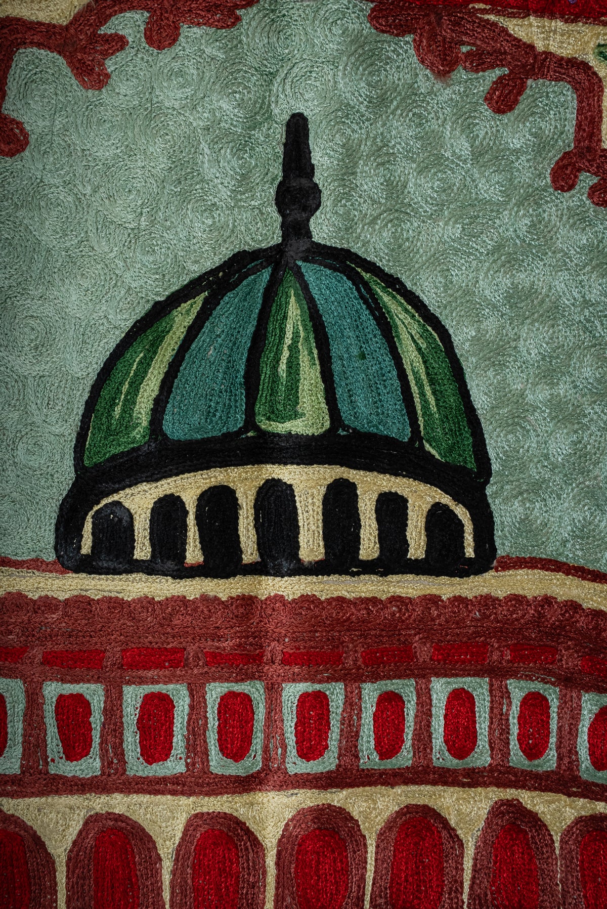 Close-up of the green dome above red windows and arches embroidered on the pale green prayer mat