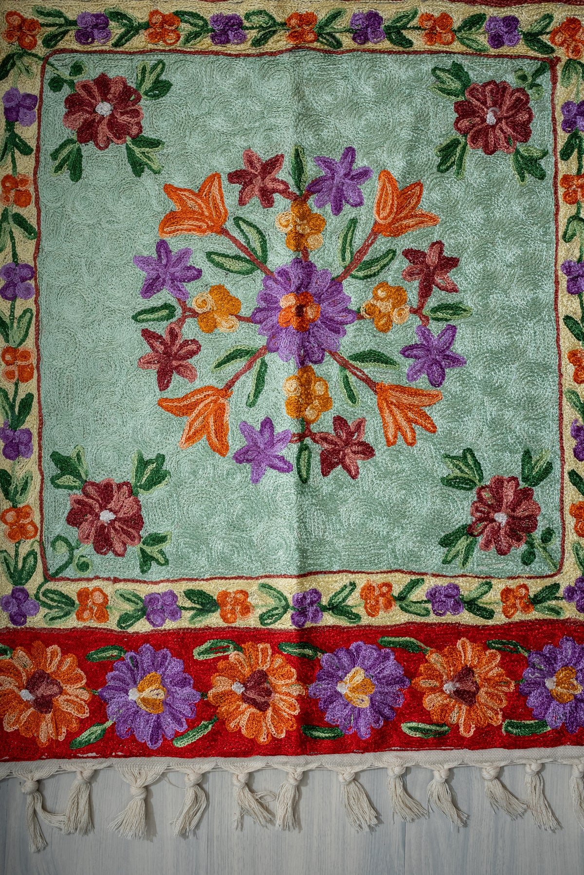 Lower section of the pale green prayer mat showing a floral circle, small corner flowers, red border and cream tassels