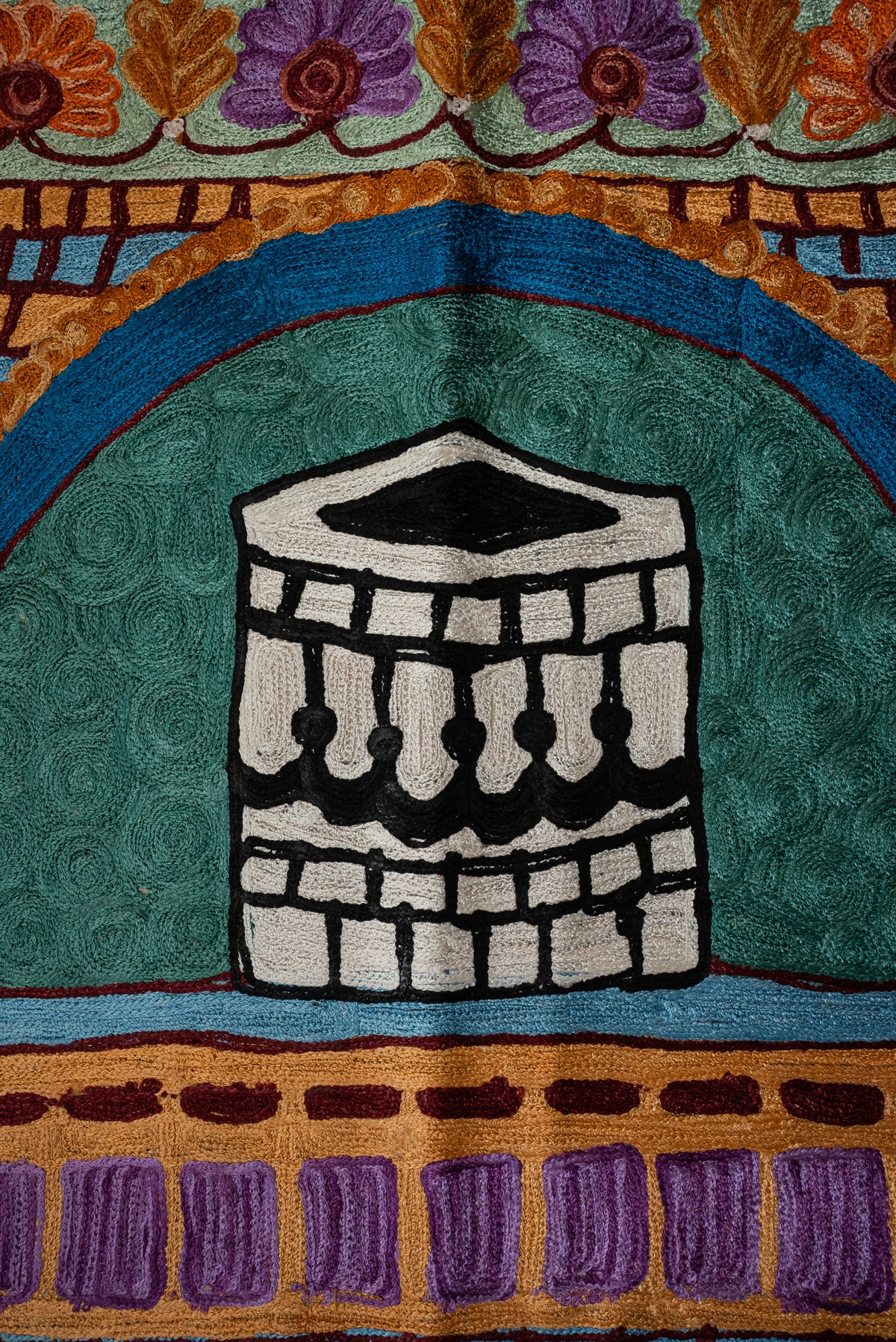 Close-up of a black-and-white embroidered Kaaba motif beneath a blue arch on a teal prayer mat