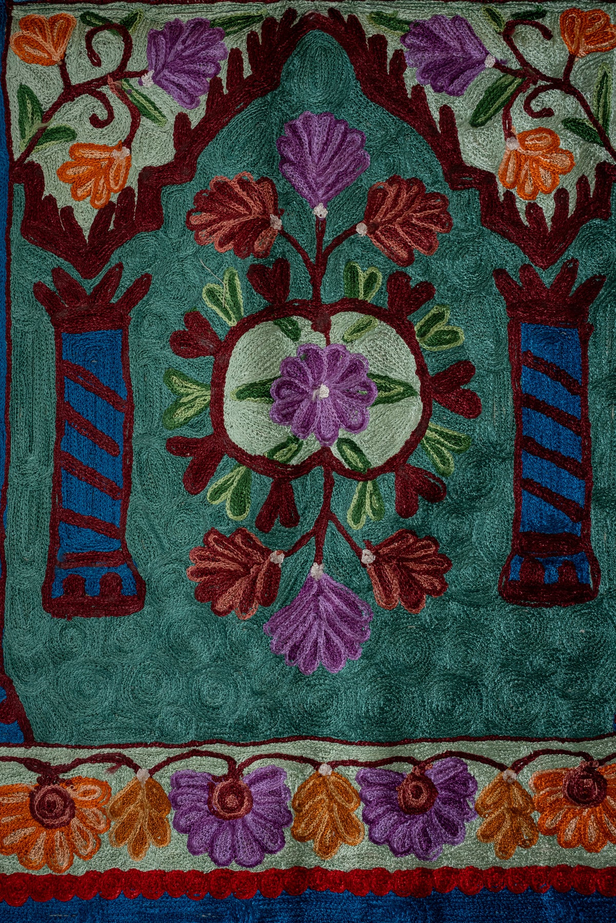 Lower section of a teal prayer mat, with a purple floral medallion beneath an arch and orange-purple flower borders