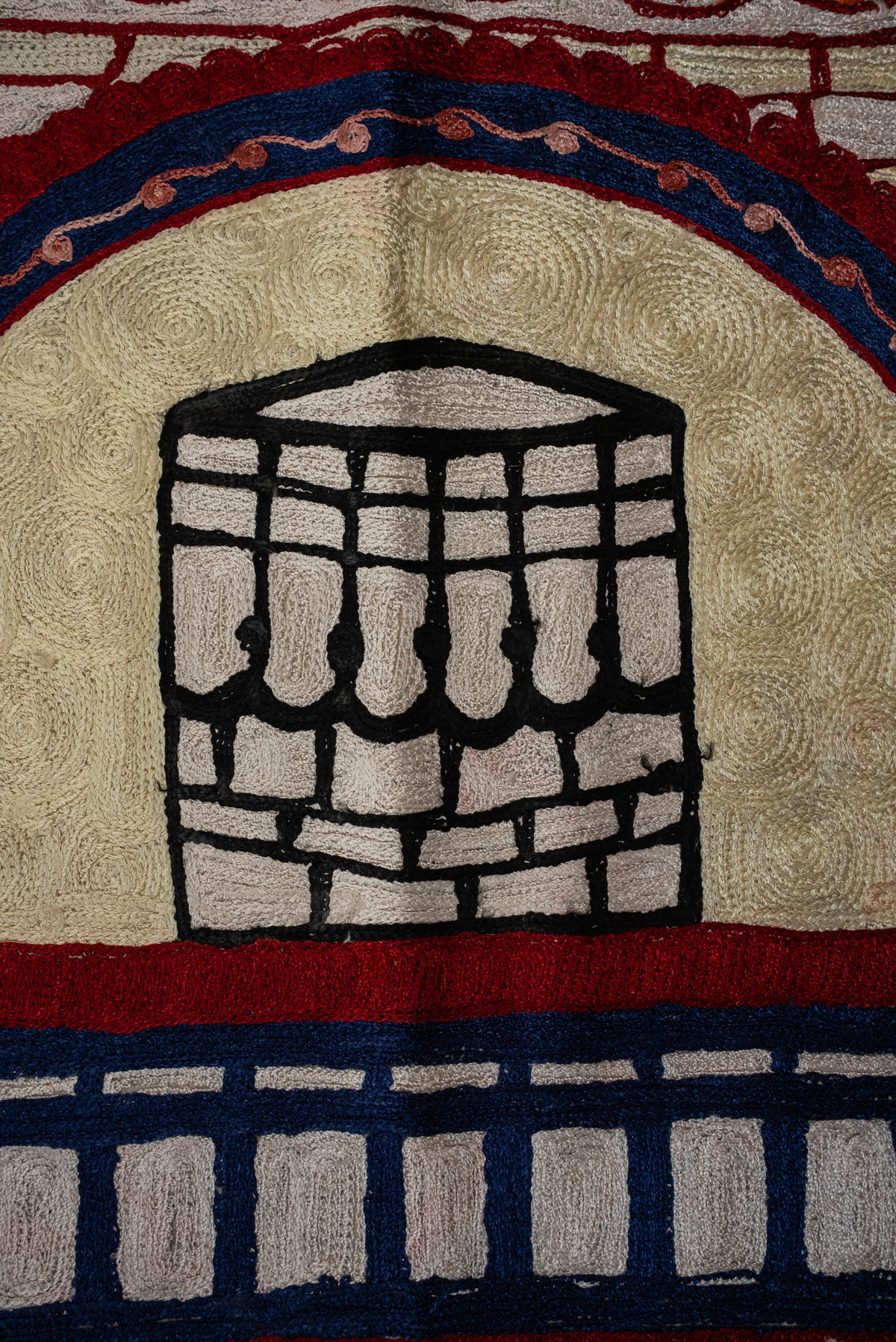 Black-and-cream embroidered Kaaba motif beneath a red and dark blue arch on the cream prayer mat