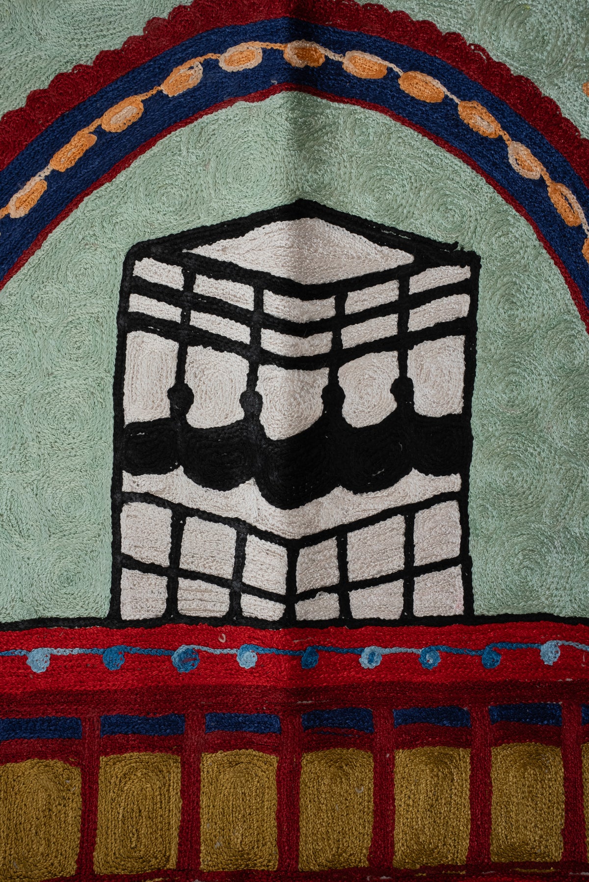 Close-up of the black-and-white Kaaba motif under a navy and red arch on the pale green prayer mat