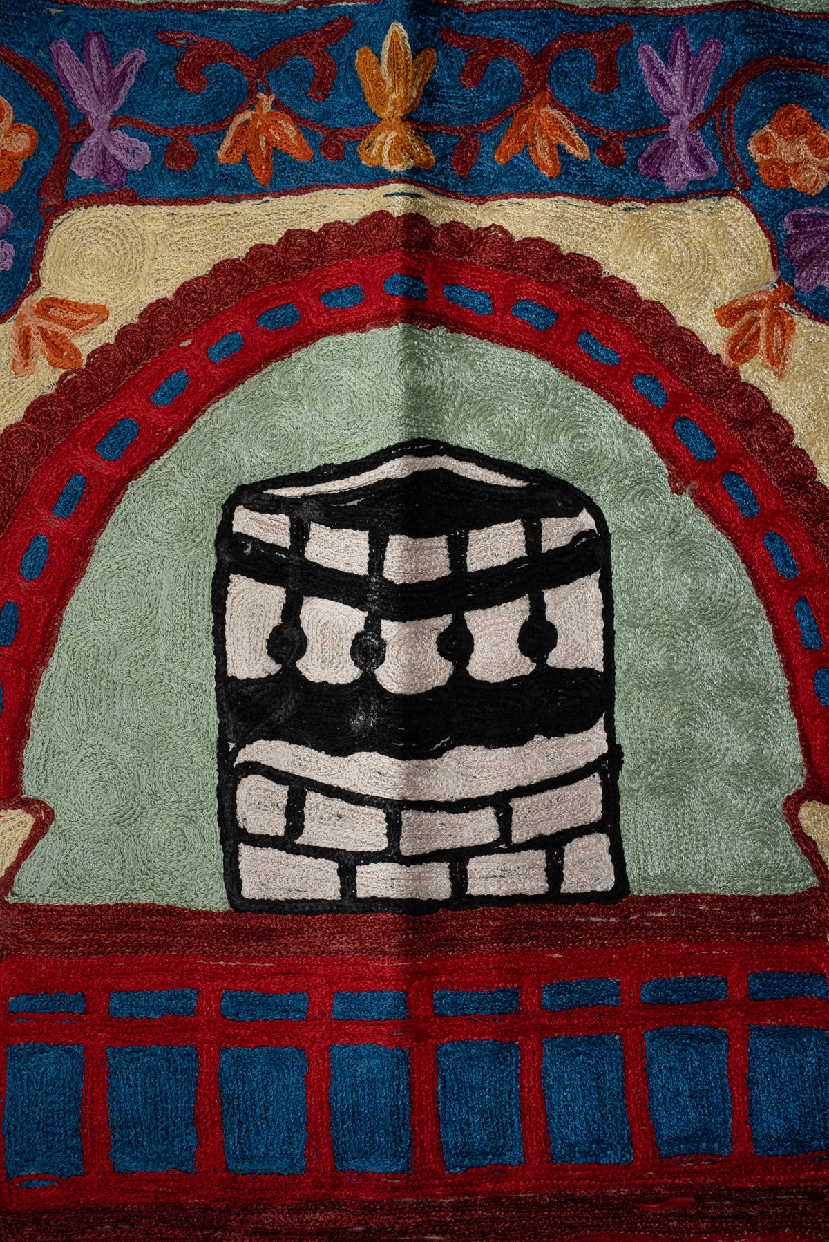 Black-and-white Kaaba motif beneath a red arch with blue accents on a pale green embroidered prayer mat