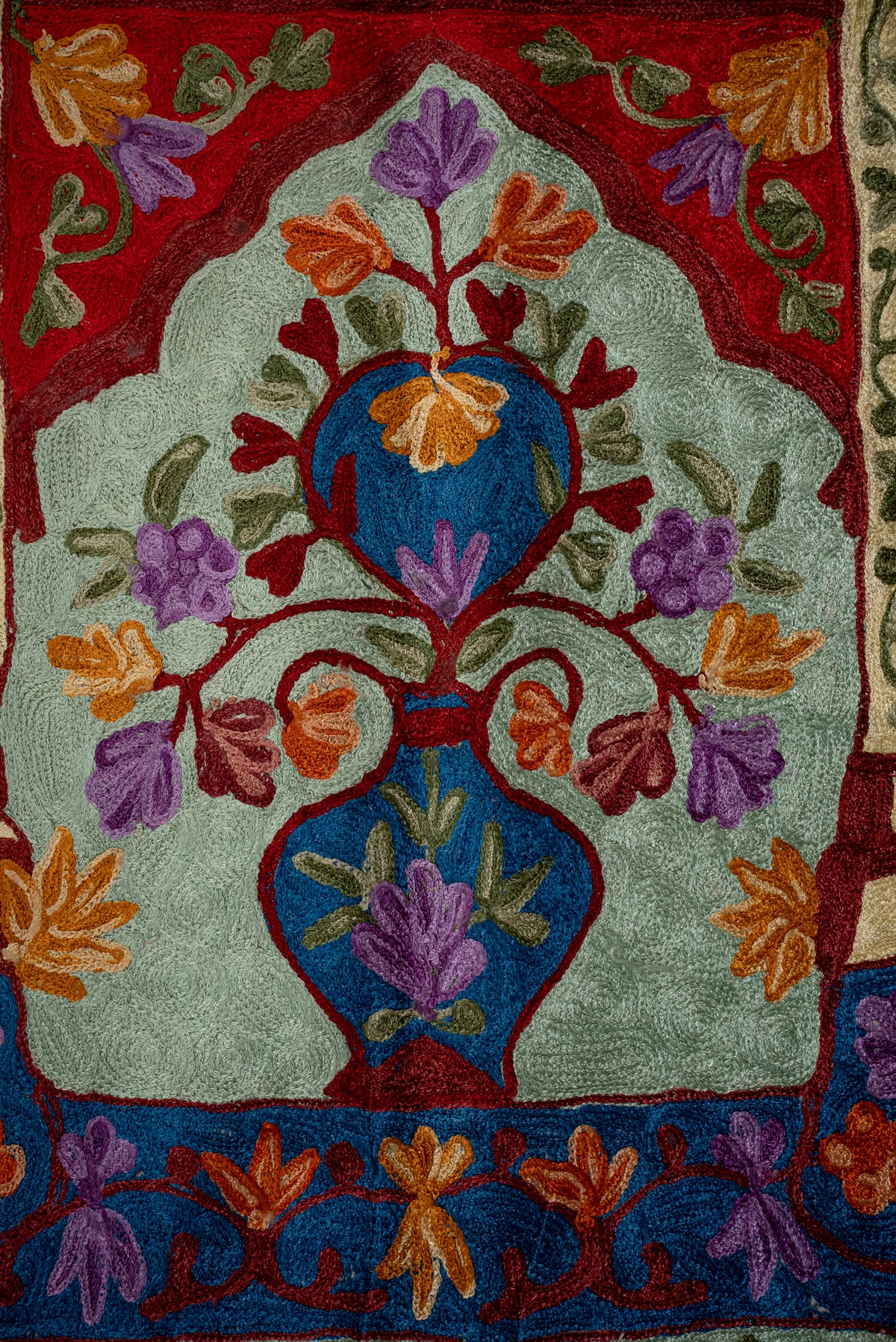 Blue vase with orange and purple flowers embroidered on the pale green centre of a prayer mat beneath a red arch