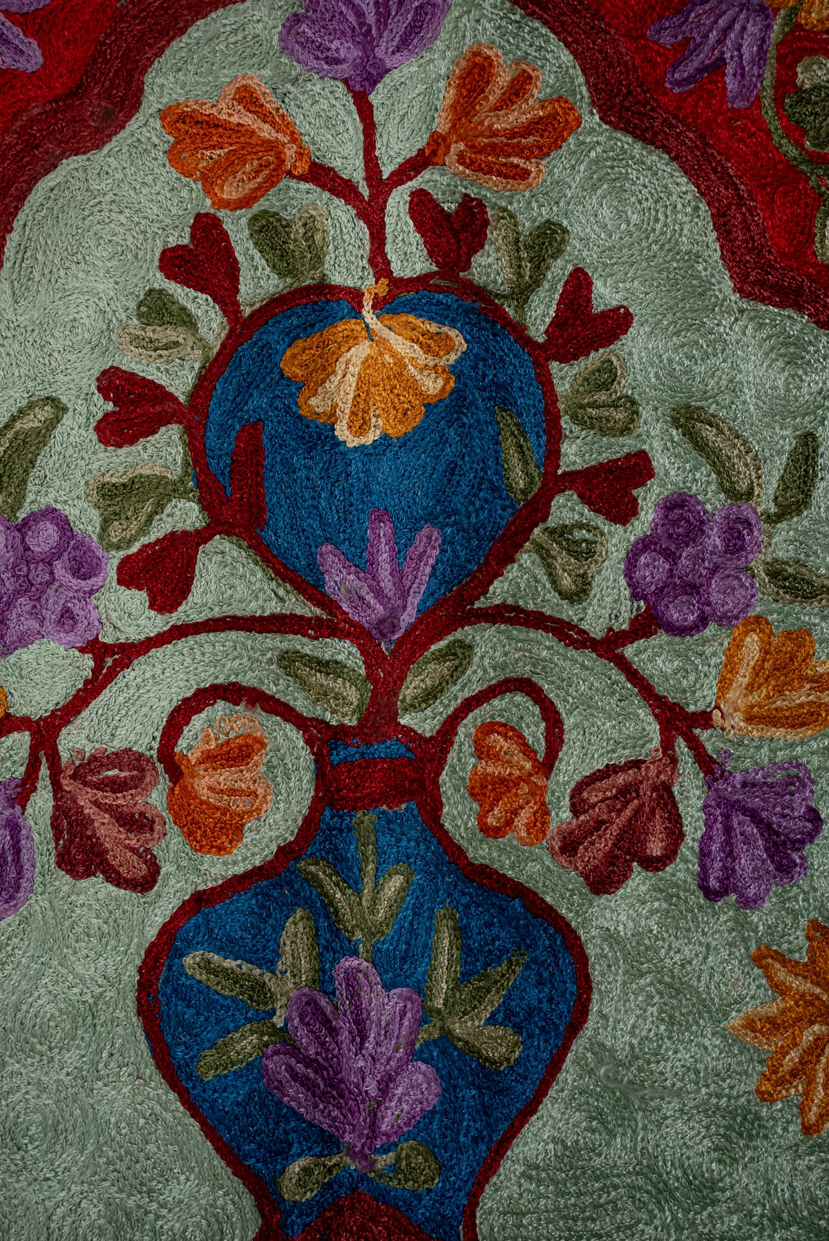 Close-up of orange and purple floral embroidery with a blue vase on the pale green centre of a prayer mat