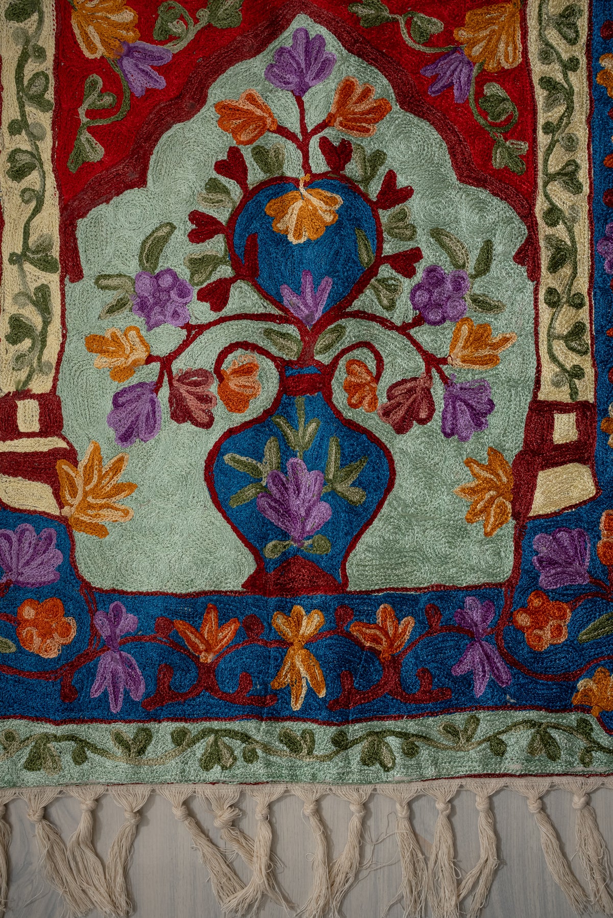 Lower section of a pale green prayer mat with a blue floral vase, blue flower borders, leafy edging and cream tassels