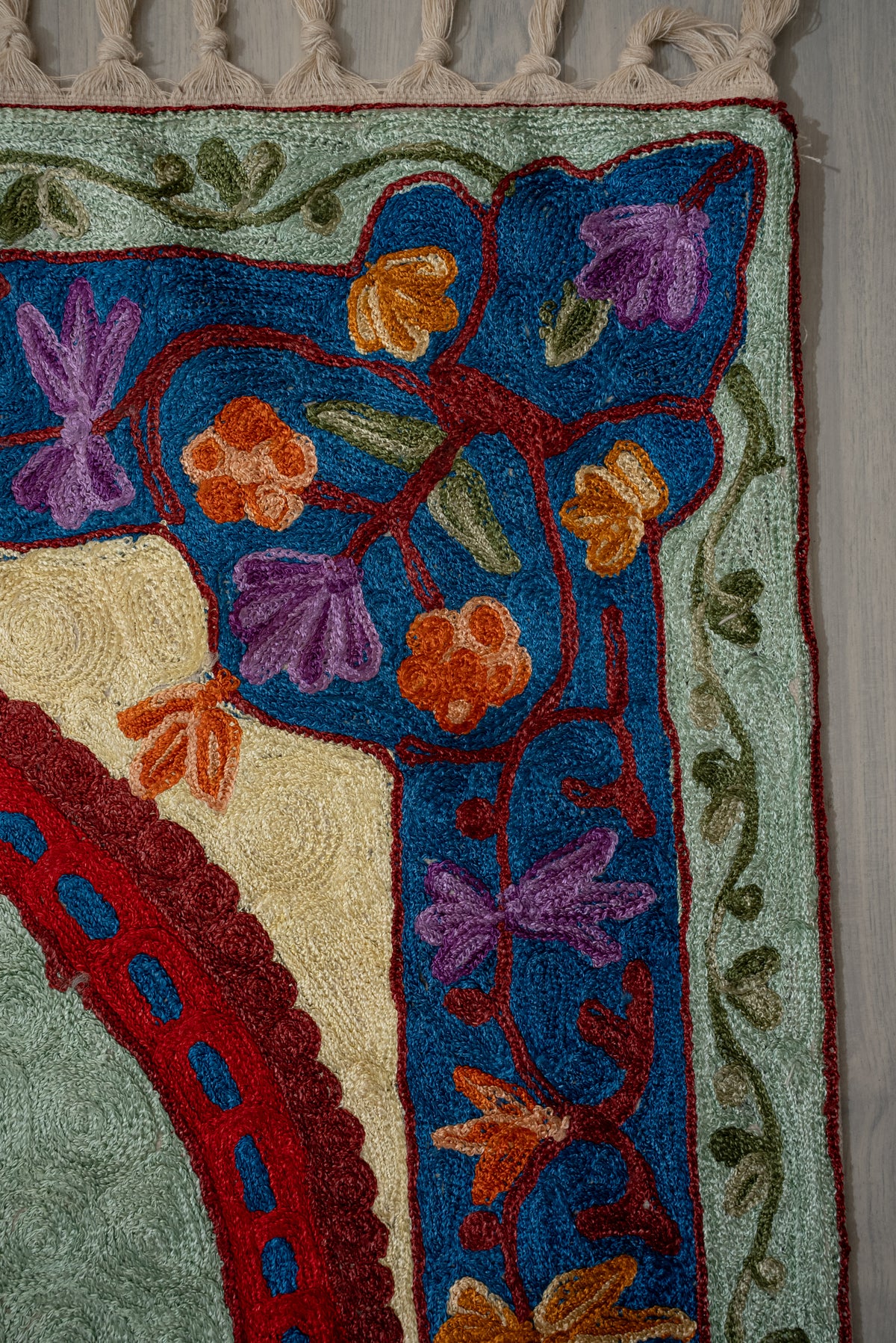 Upper corner of an embroidered prayer mat with orange and purple flowers on blue, pale green leafy edging and cream fringe