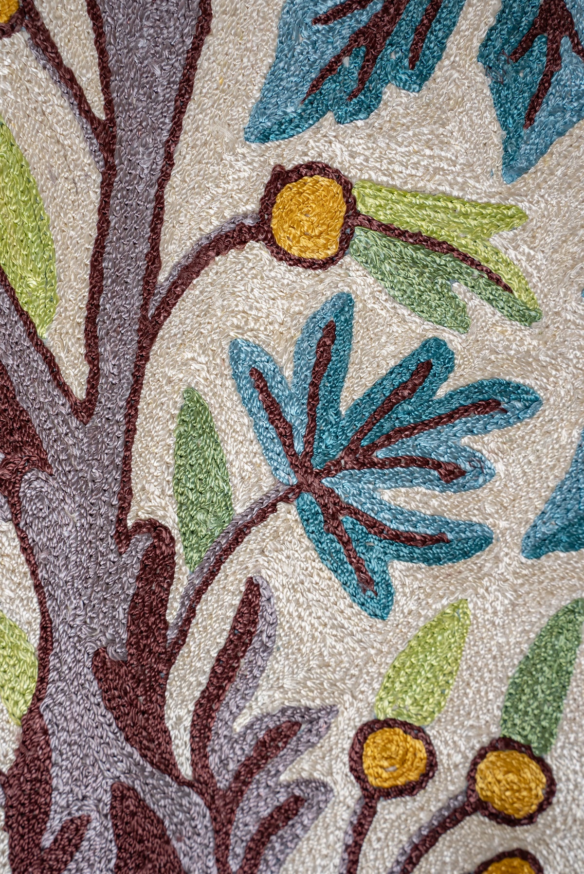 Close-up of a teal leaf beside a grey-brown tree trunk and golden berries on an ivory chain-stitch rug