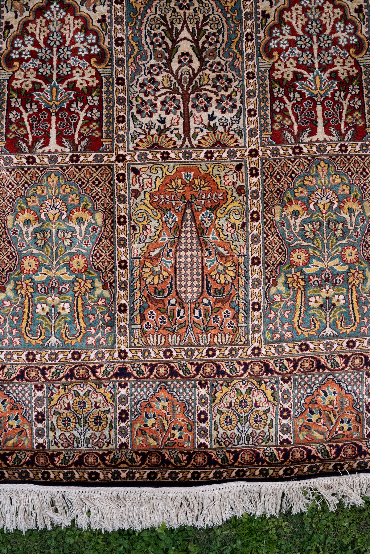 End section of a silk carpet with rows of red, teal and ivory floral panels above patterned borders and cream fringe