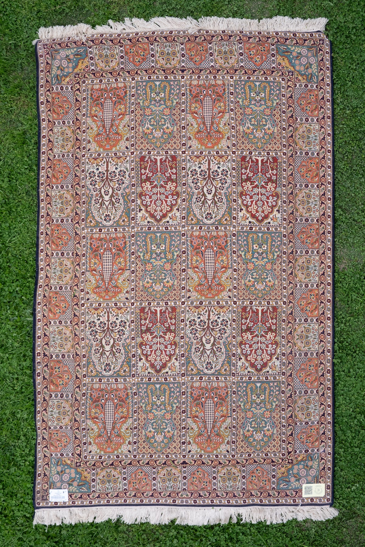 Silk carpet laid flat on grass, with a grid of multicolour tree and floral panels, cream fringe and corner labels