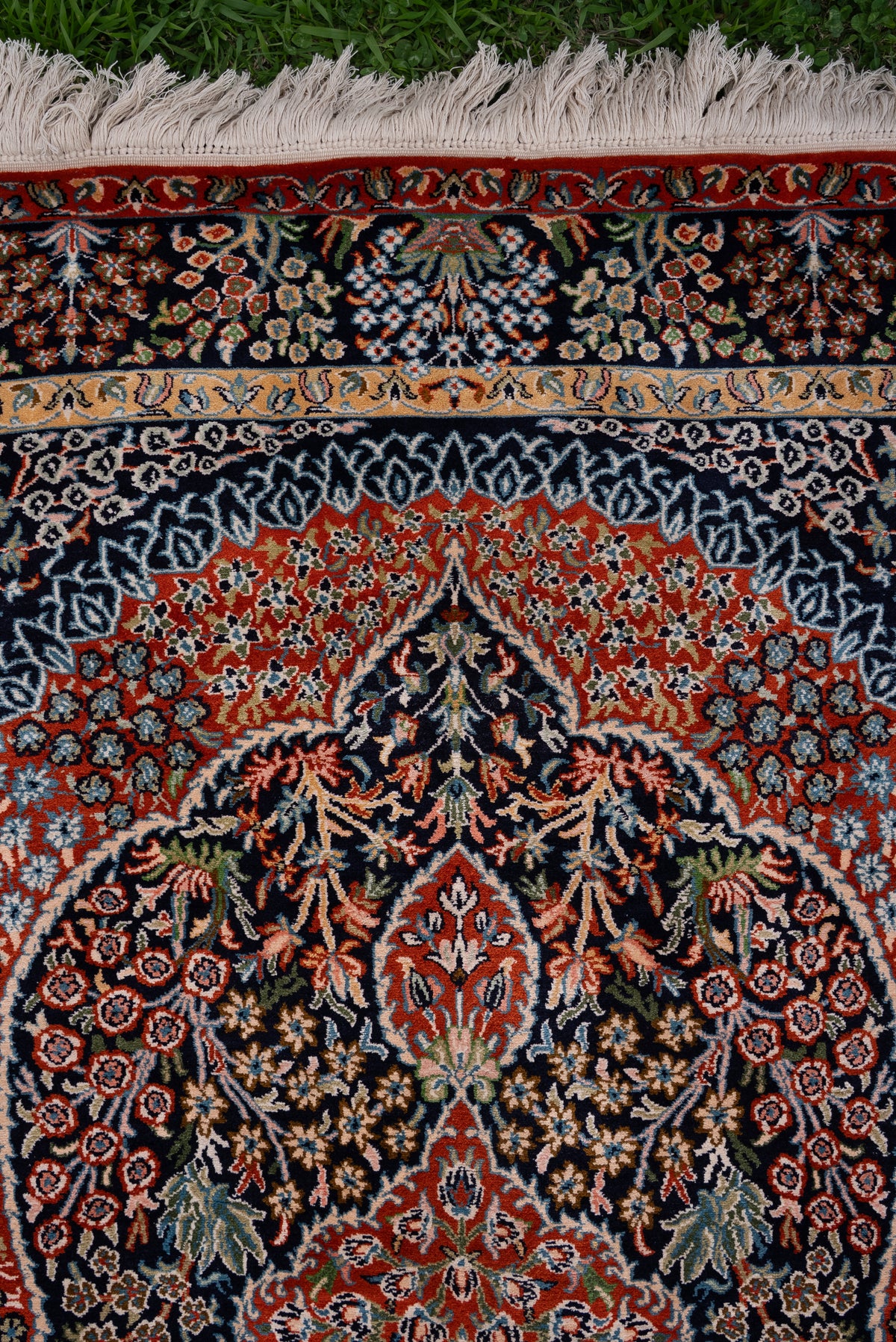 Upper end of the dark silk carpet with red arches, floral vines, patterned borders and pale fringe