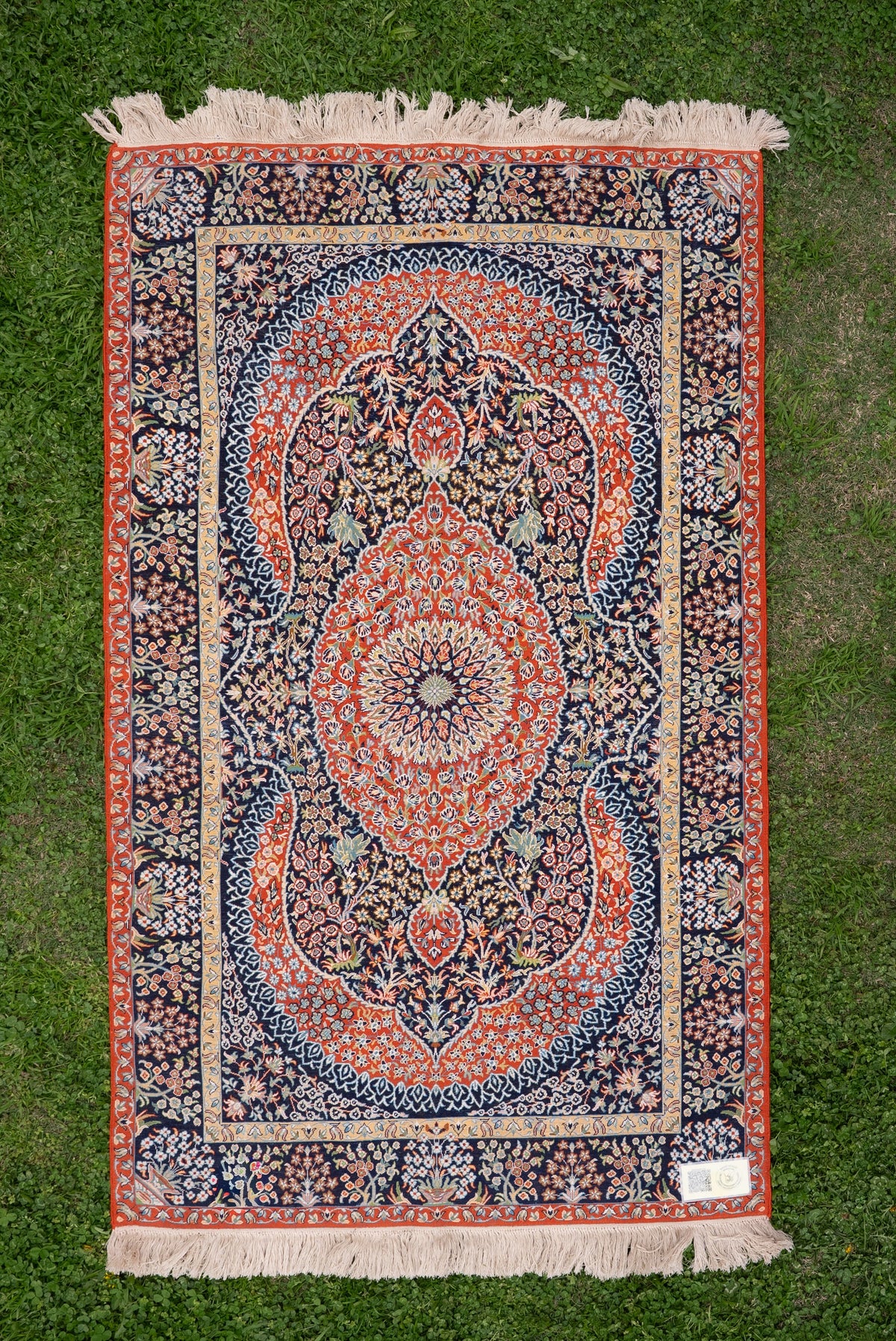 Dark silk carpet laid flat on grass, showing a red central medallion, floral borders, pale fringe and corner label
