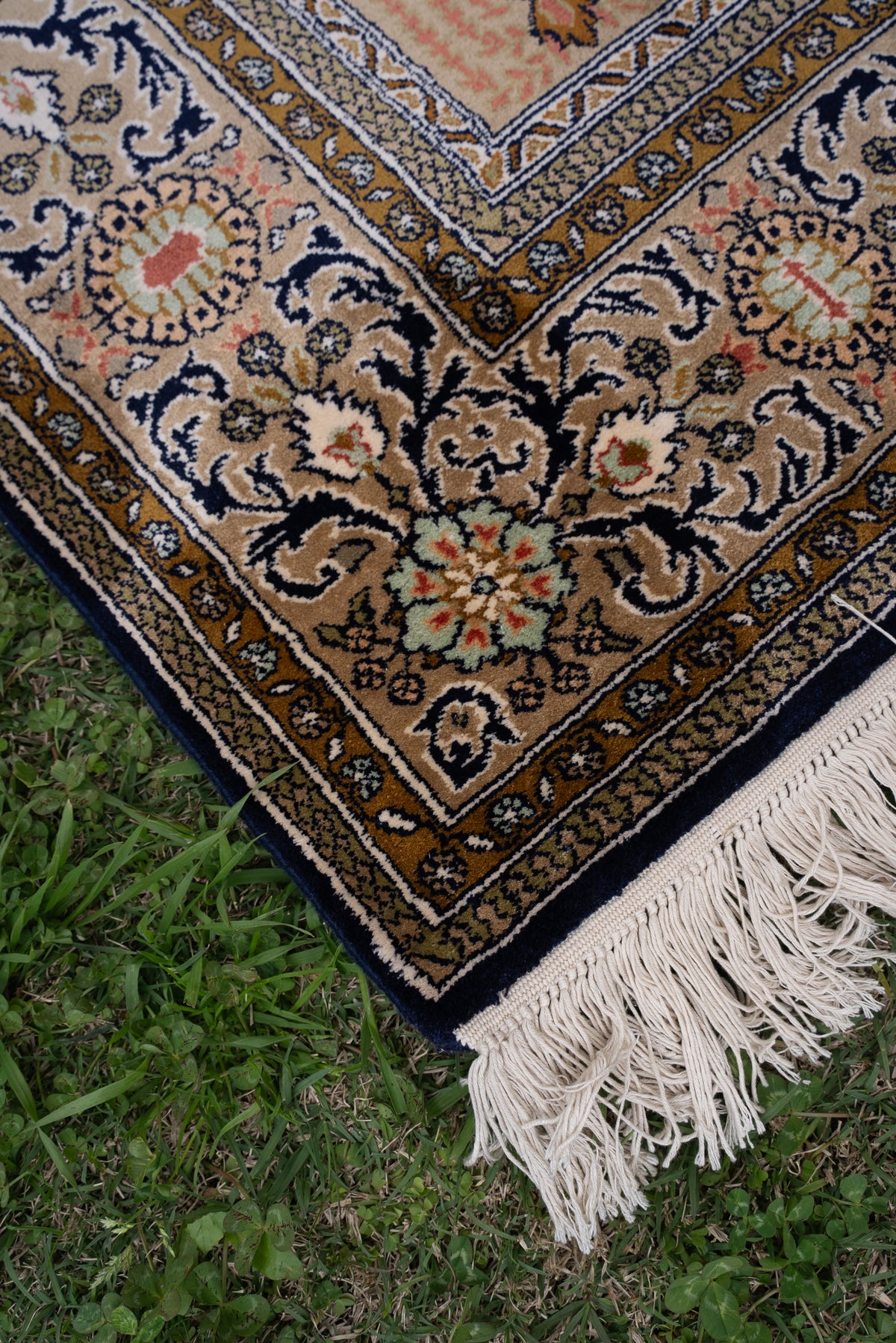 Angled corner of a silk carpet on grass, with navy floral vines, gold borders and white fringe