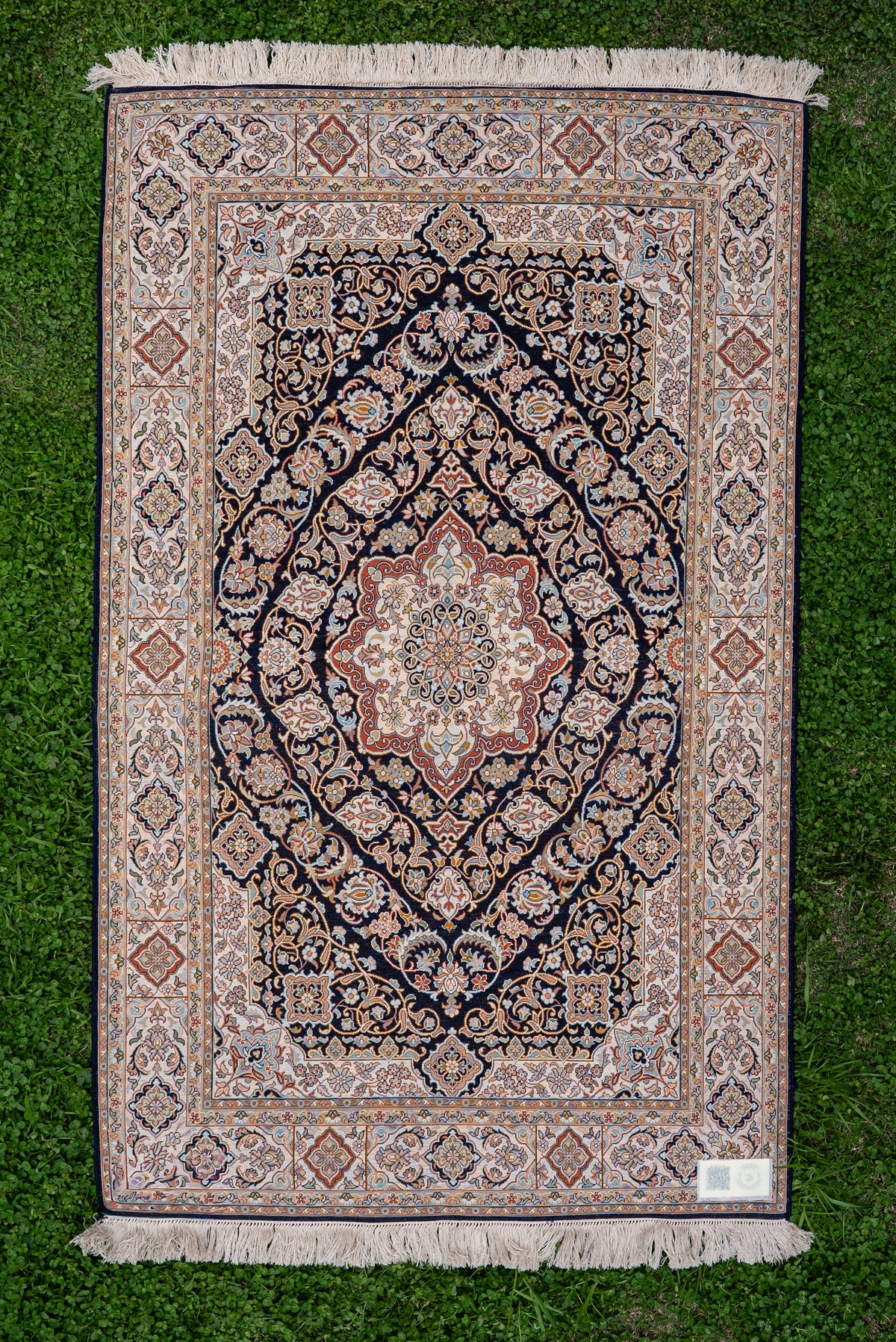 Reverse of a dark floral carpet laid on grass, showing its central medallion, pale layered borders, corner label and ivory fringe