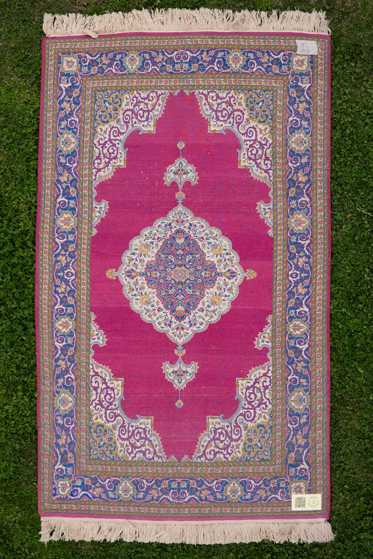 Reverse of the rose-pink medallion carpet laid flat on grass, with corner labels and ivory fringe