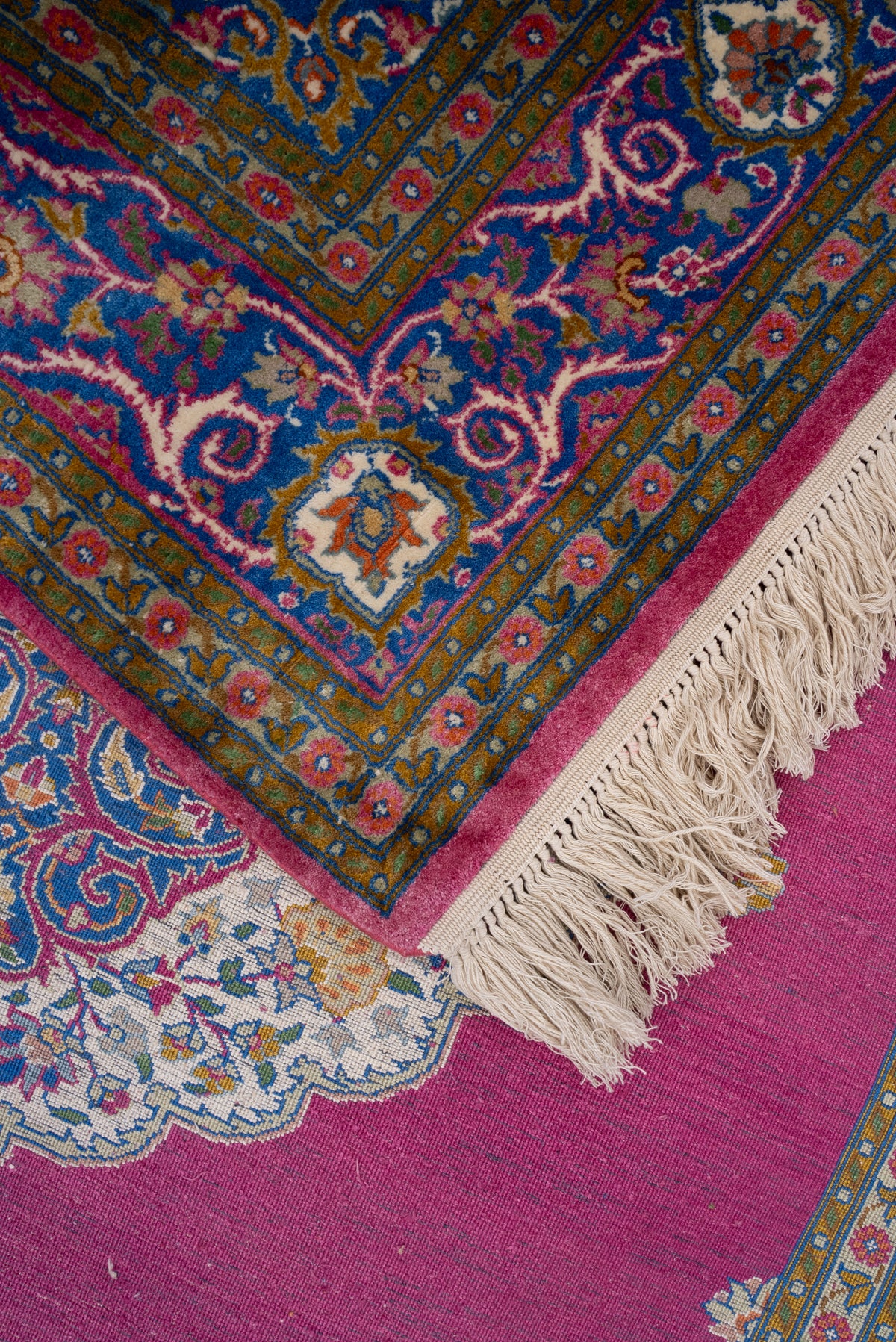 Folded carpet corner showing its blue floral border and ivory fringe over the rose-pink field