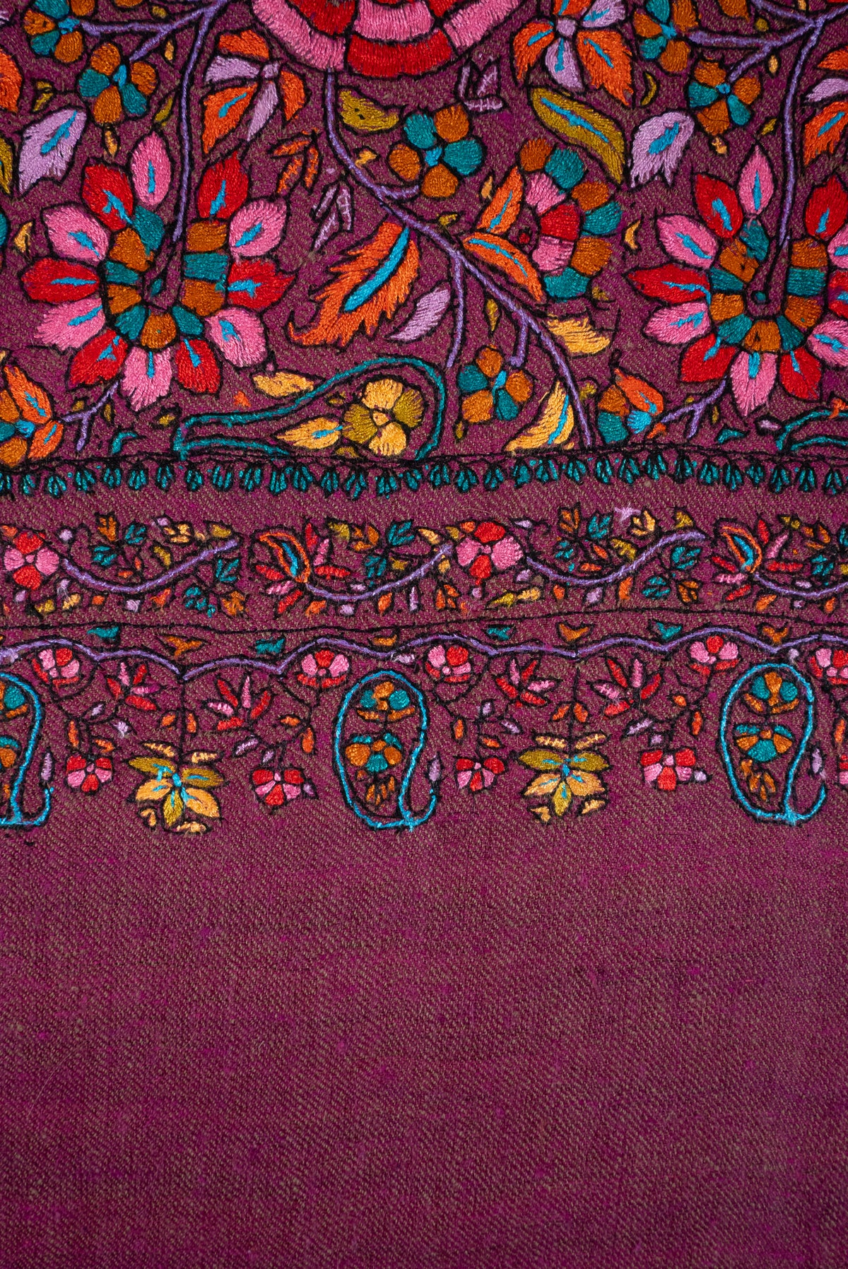 Macro view of a plum Pashmina shawl border, with colourful flowers, curling vines and turquoise-outlined hanging paisleys