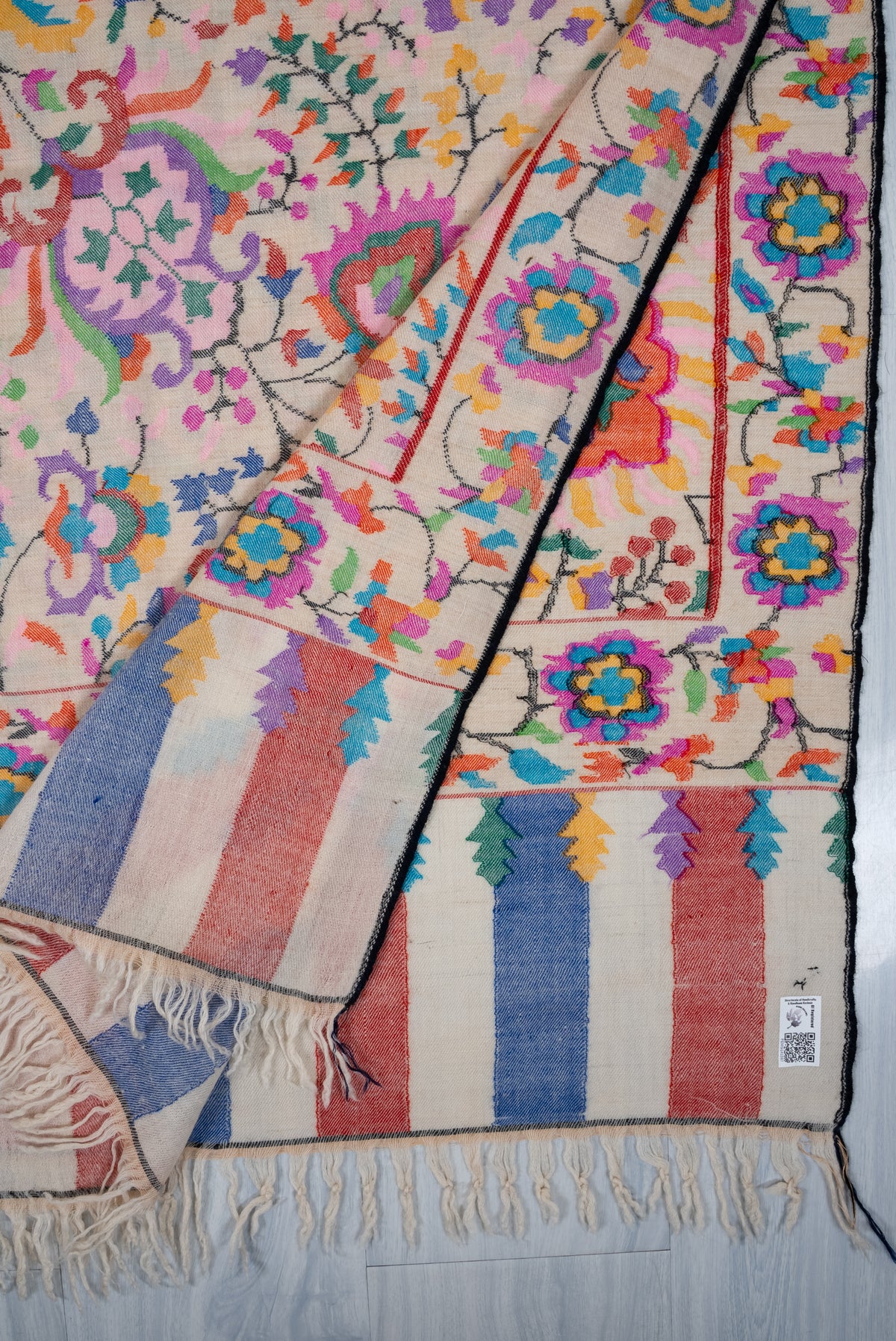 Folded corner of ivory Pashmina shawl with colourful floral borders, striped end panels, a small label and cream fringe