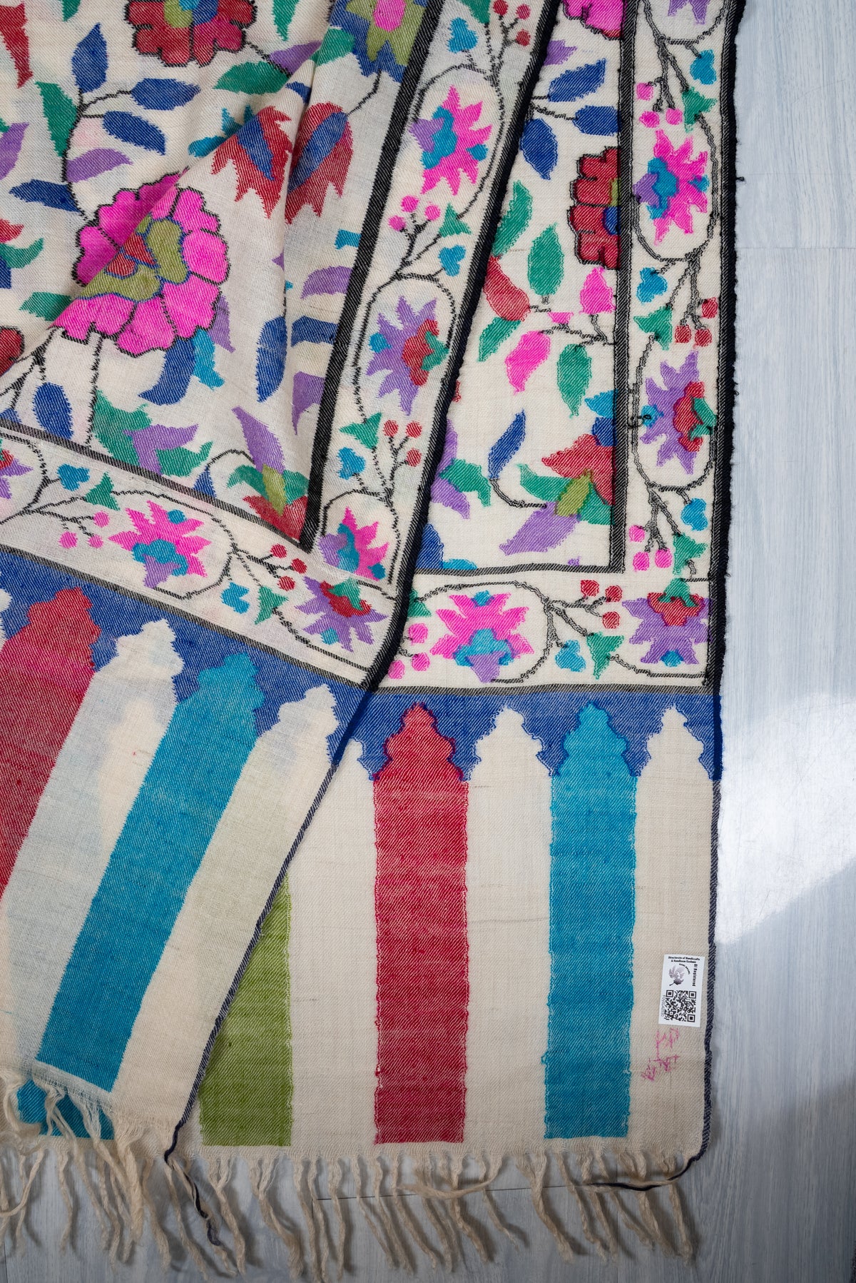 Folded corner of an ivory Pashmina shawl showing colourful flowers, striped end panels, cream fringe and a small label