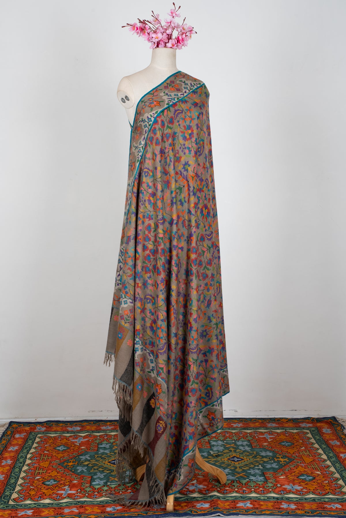 Full-length taupe Pashmina shawl draped on a mannequin, with colourful floral patterns and striped fringed ends