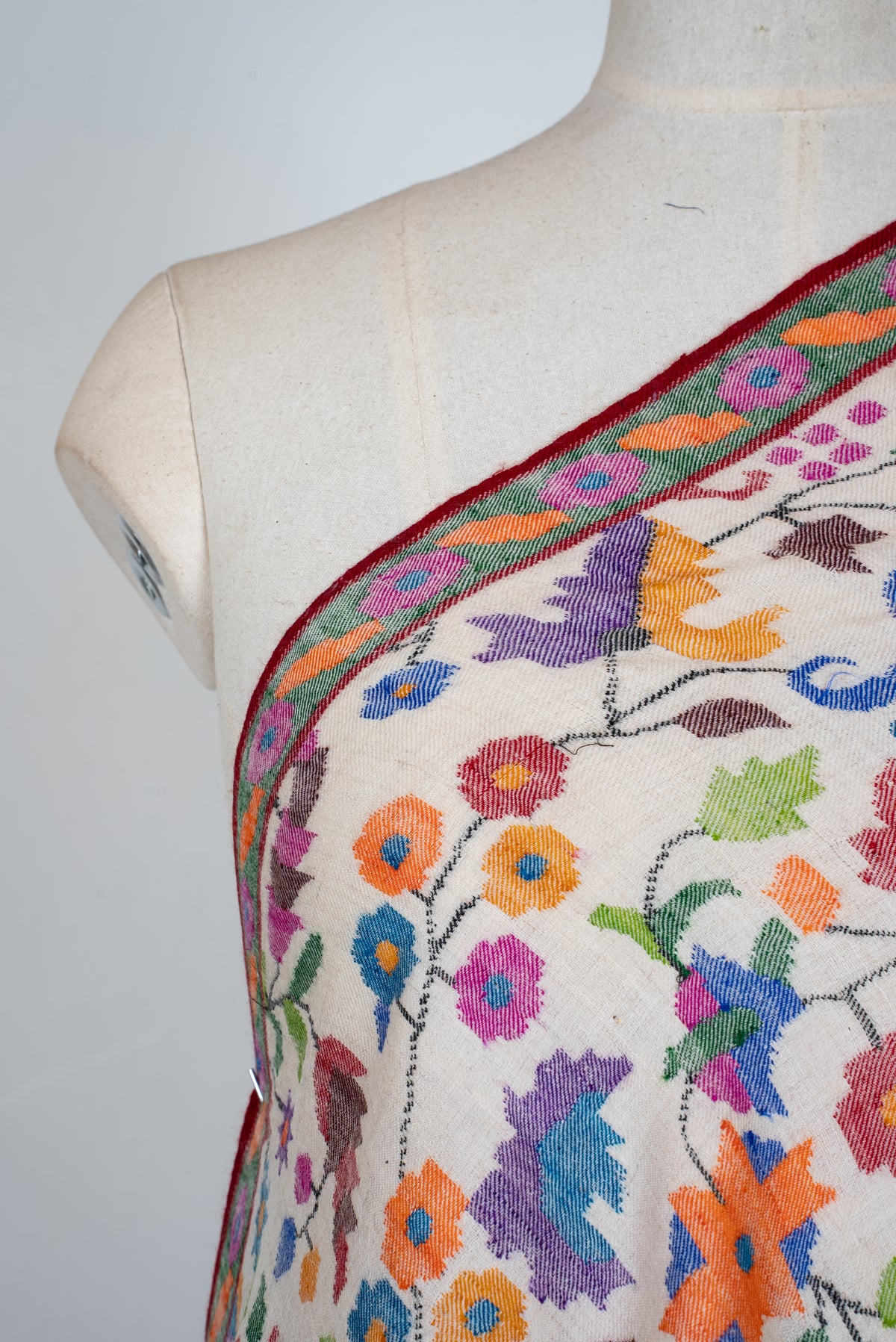 Close-up of an ivory Pashmina shawl across a mannequin's chest, with orange, blue, purple and pink flowers on fine dark vines.