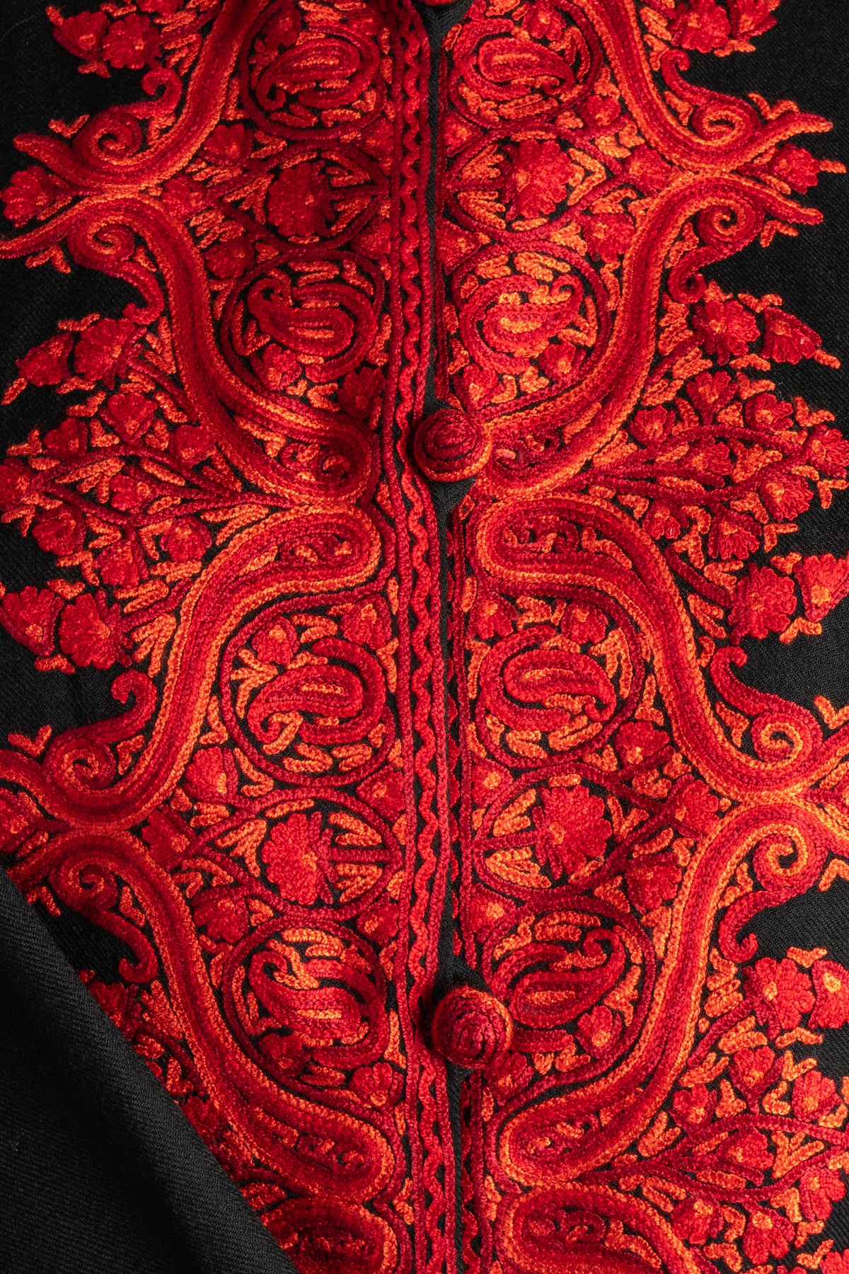 Close-up of raised red Aari paisleys and small flowers outlined in orange beside red covered buttons on a black jacket