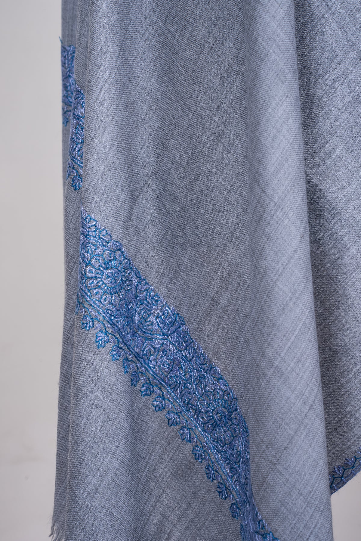 Detail of a draped powder-blue merino wool scarf, with a blue embroidered floral band across the end and fine edging.