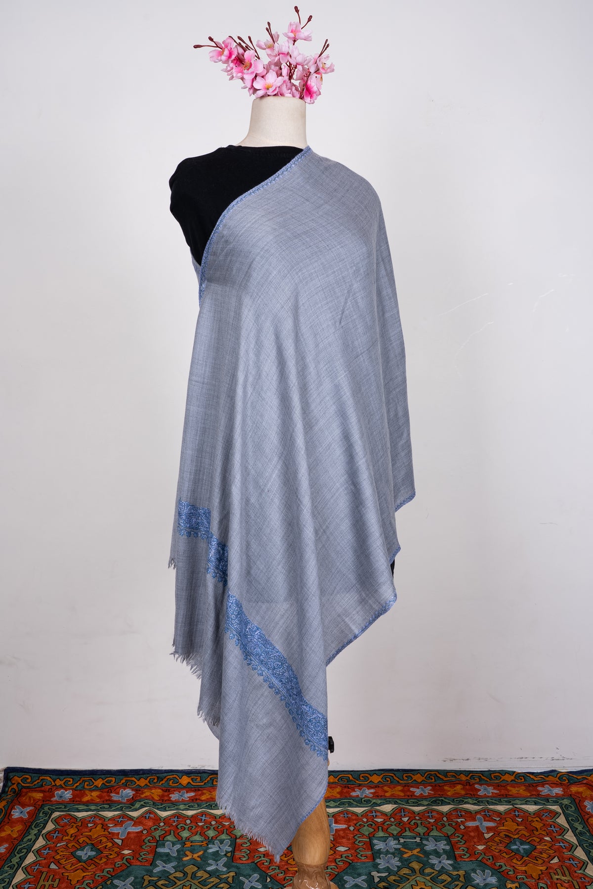 Powder-blue merino wool scarf draped on a mannequin, with blue embroidered edging and a wider floral band near the fringed end.