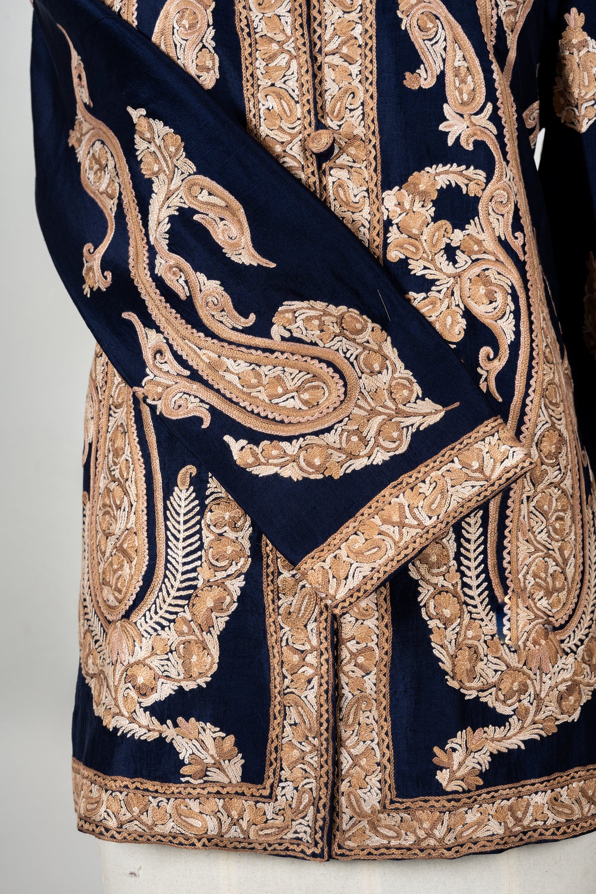 Detail of beige Aari paisley embroidery across a navy jacket sleeve, with dense floral borders at the cuff, front and hem