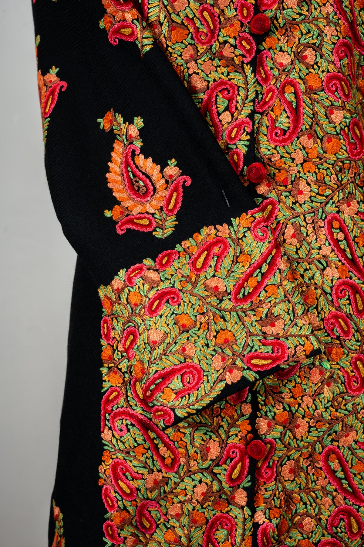 Close-up of the black jacket’s folded cuff and front panel, densely embroidered with red paisleys, orange flowers and green leaves.