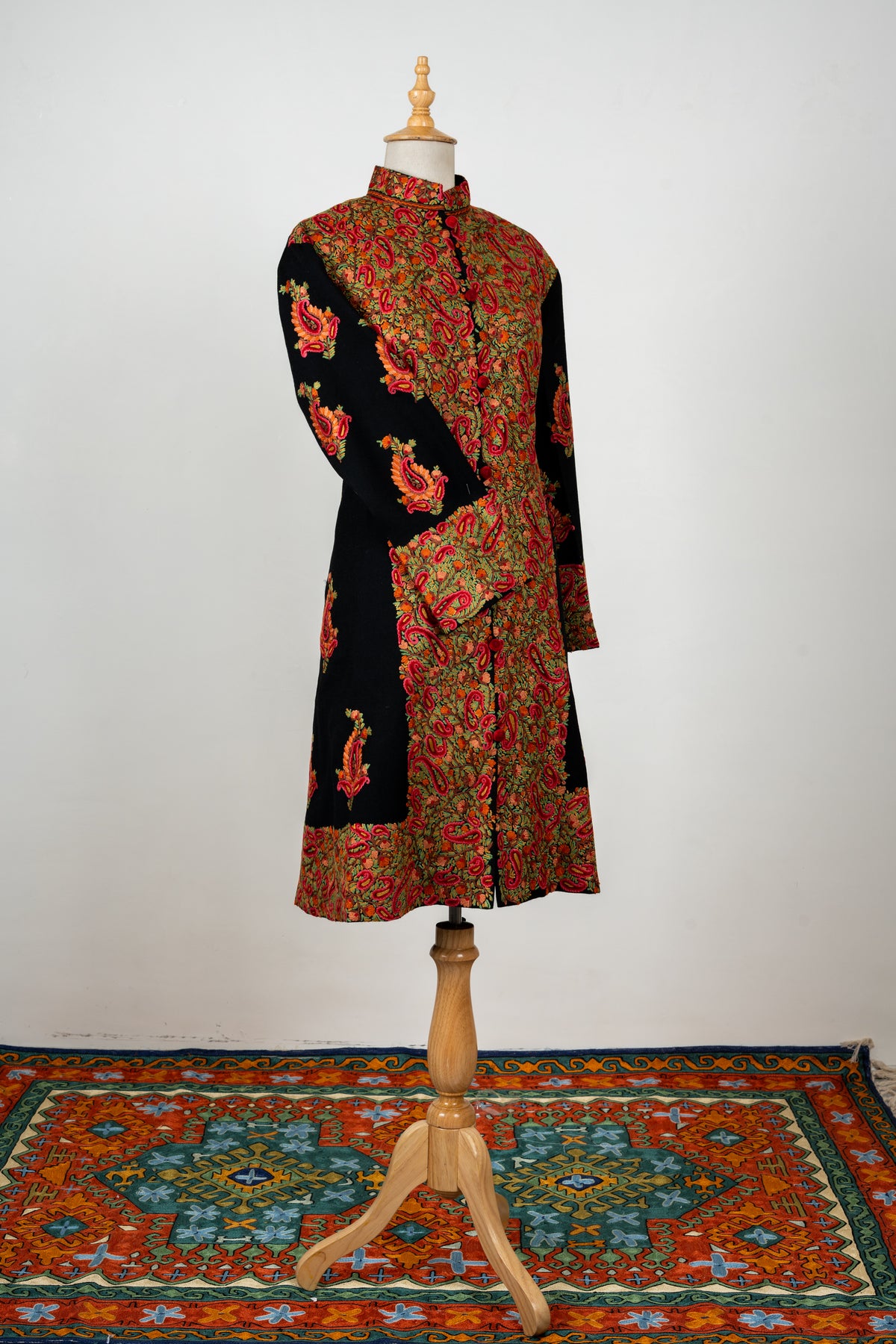 Angled full front of the black long jacket with broad multicoloured paisley embroidery and one sleeve crossed.