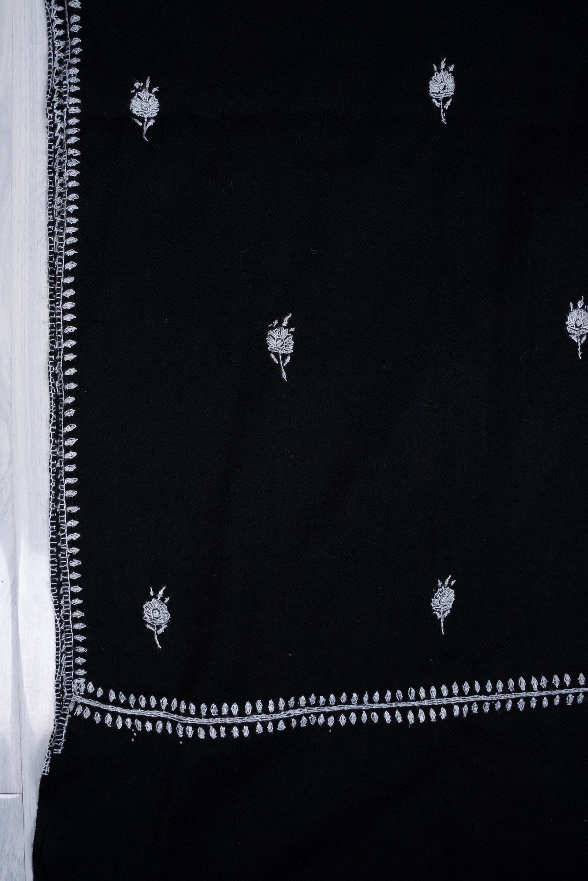 Corner of a black scarf laid flat, with small white embroidered flower sprigs inside intersecting fine leafy borders.