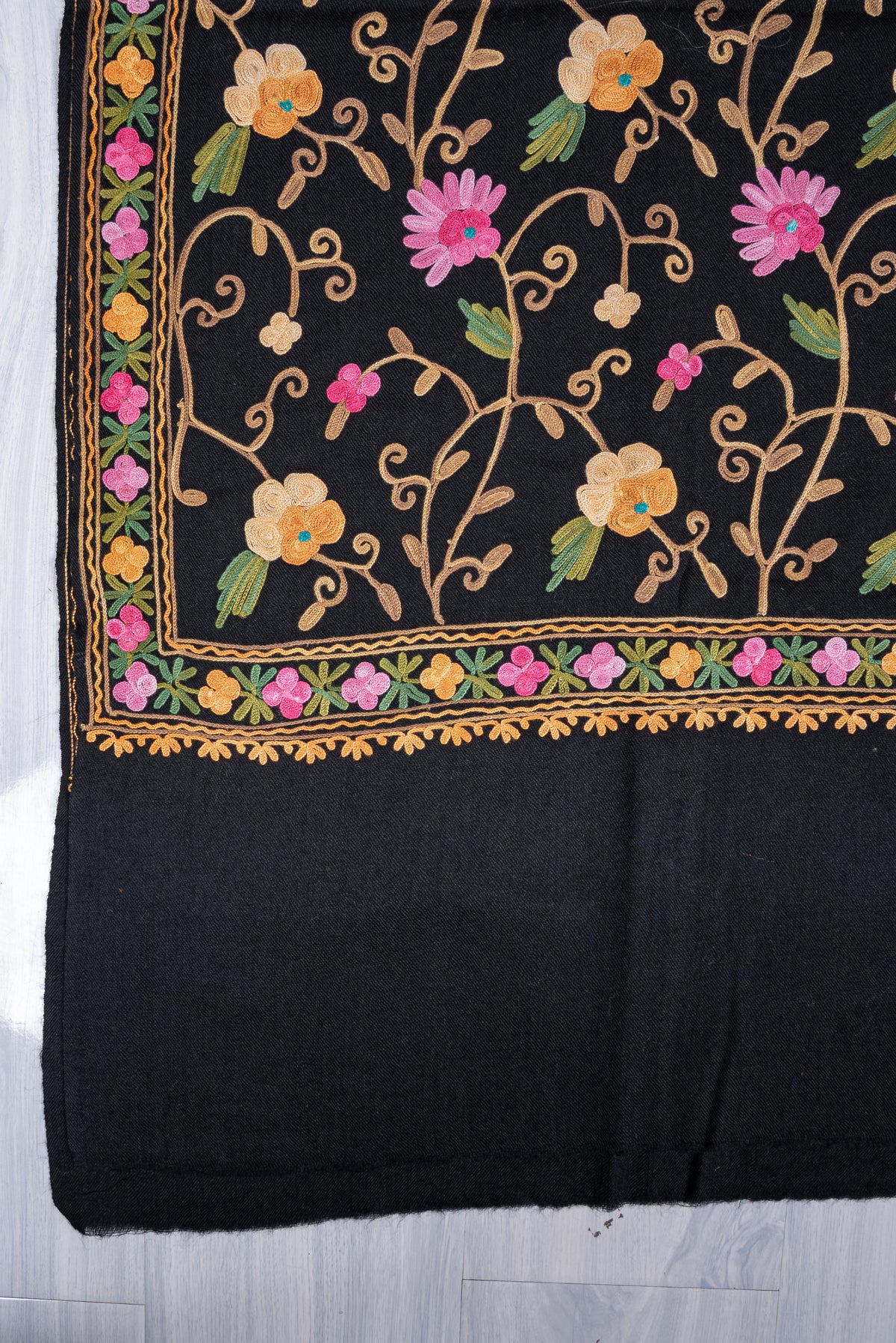 Corner of a black shawl laid flat, with pink and cream Aari flowers framed by a narrow floral border above a plain end.