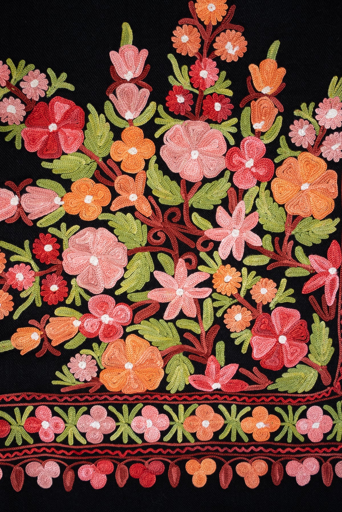 Close-up of pink, red and orange Aari flowers with green leaves above a narrow floral border on black shawl