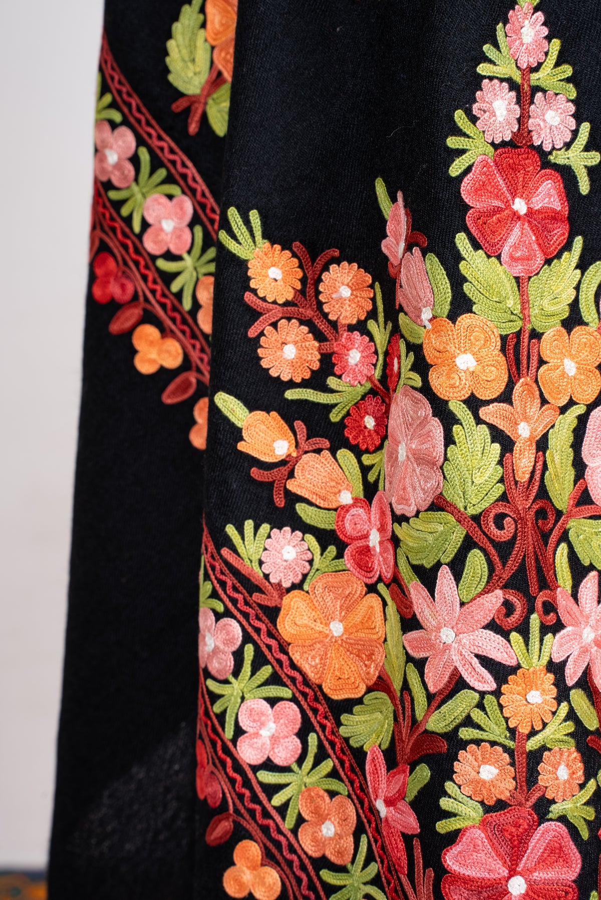 Close-up of black shawl folds with pink, red and orange Aari flowers and bright green leaves beside a floral border