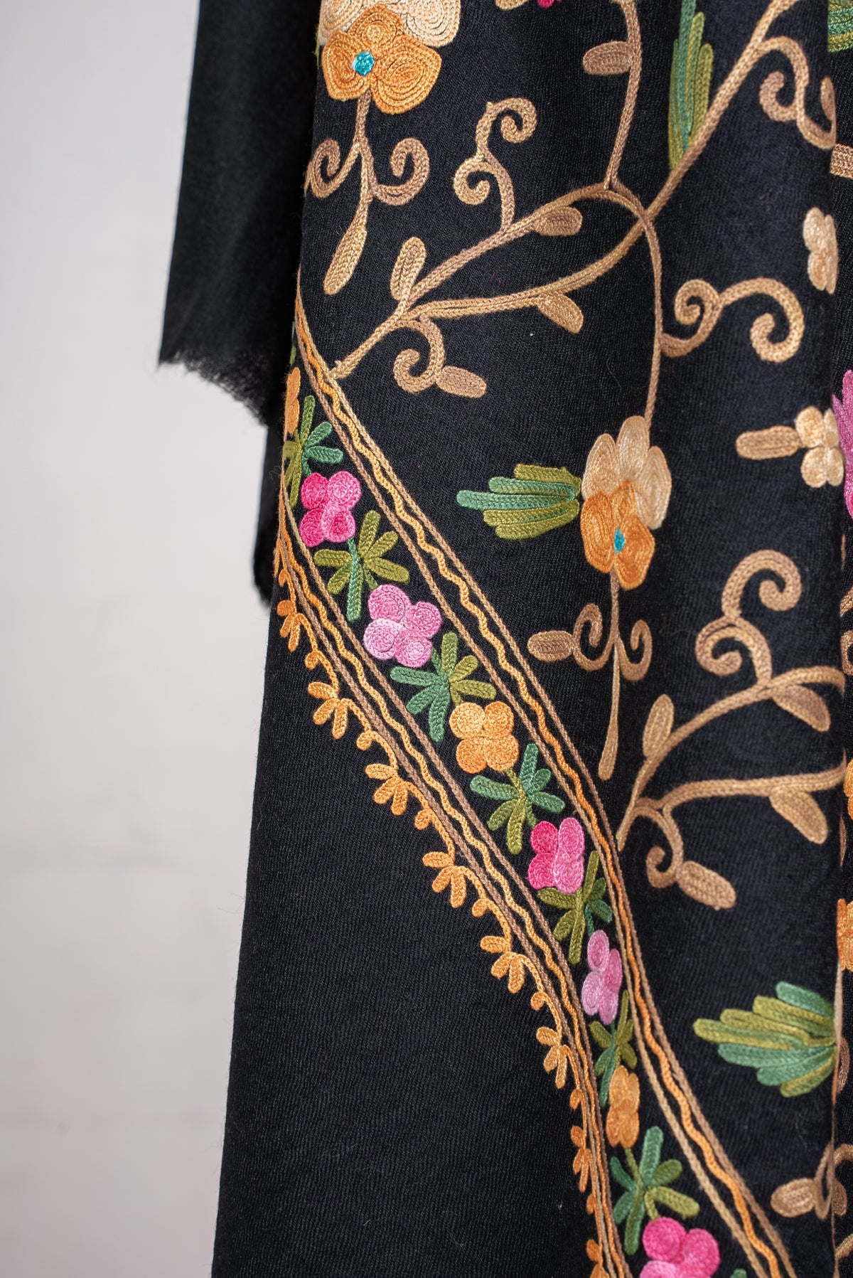 Detail of a draped black shawl, with a pink-and-green floral border beside tan Aari vines and cream flowers.