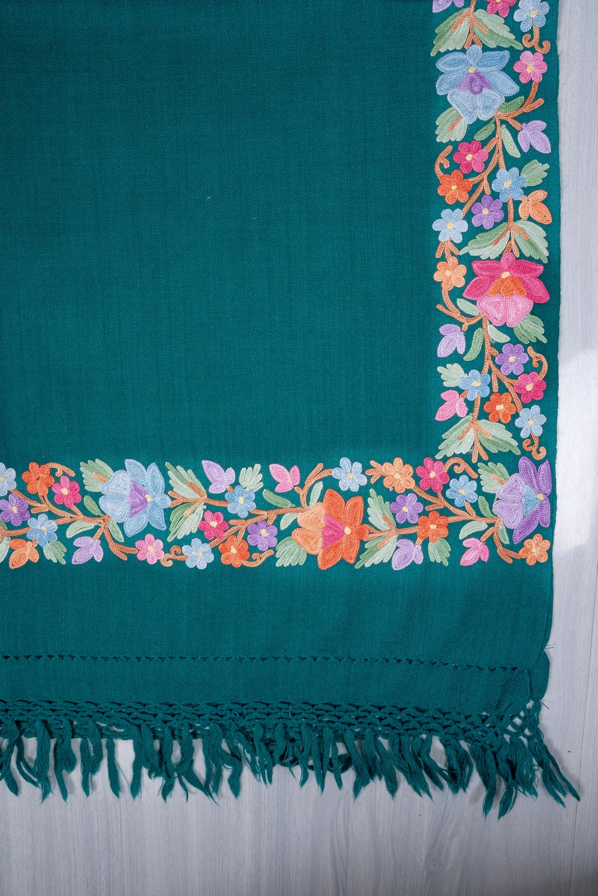 Corner of a teal shawl laid flat, showing pastel Aari floral borders meeting above a plain band and matching knotted fringe.