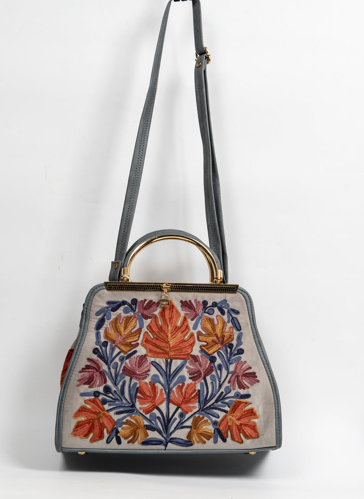 Beige embroidered handbag hanging from a grey shoulder strap, with an arched gold-toned handle and colourful leaf panel