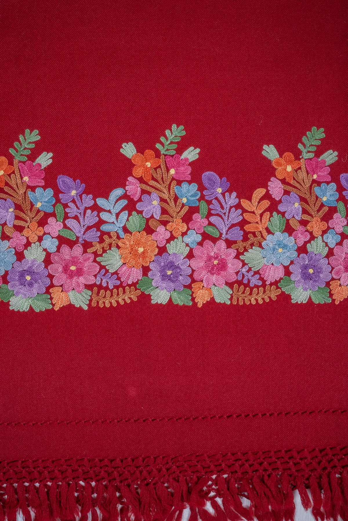 Close-up of the maroon shawl's Aari floral border, with pink, lavender, peach and blue flowers above knotted red fringe.