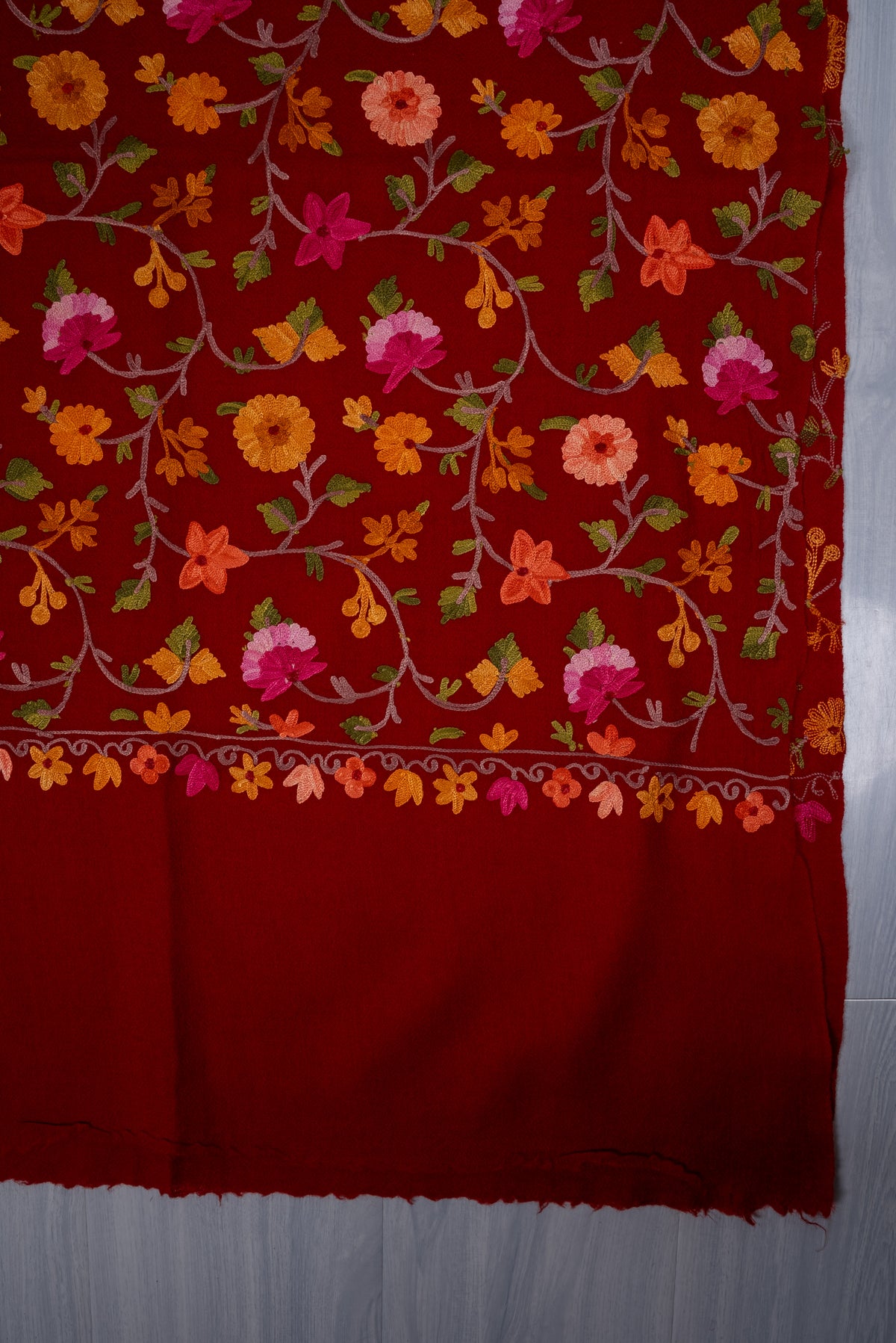Flat corner detail of the crimson shawl, showing colourful Aari flowers and a small floral border beside the plain red end