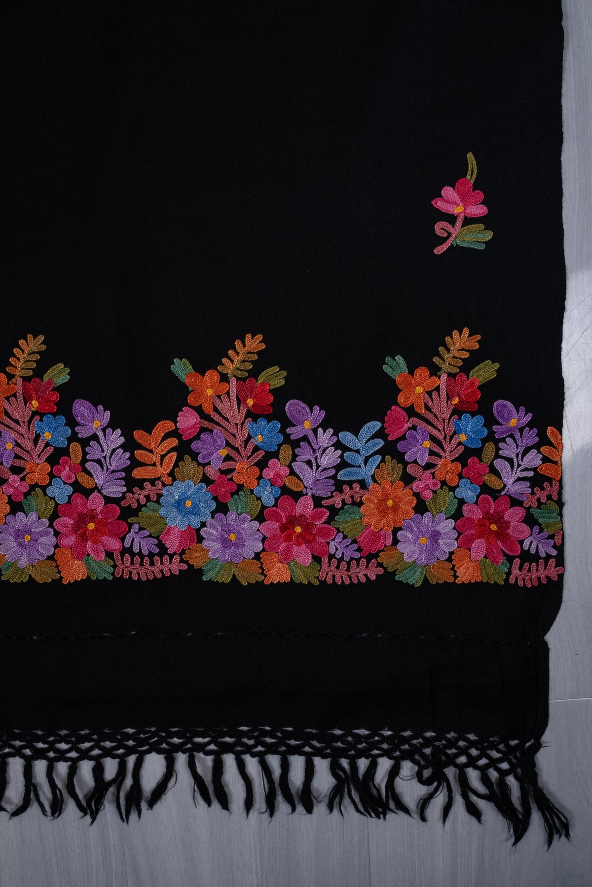 Detail of a black shawl corner laid flat, with a pink sprig above a colourful Aari floral border and black knotted fringe.