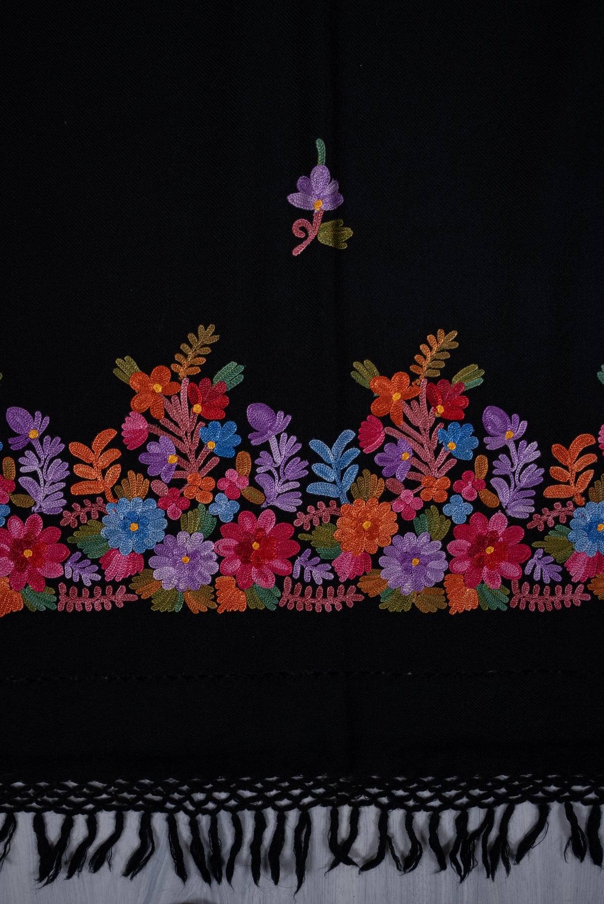 Black shawl laid flat, showing a multicoloured Aari floral border, one small purple sprig and knotted fringe.