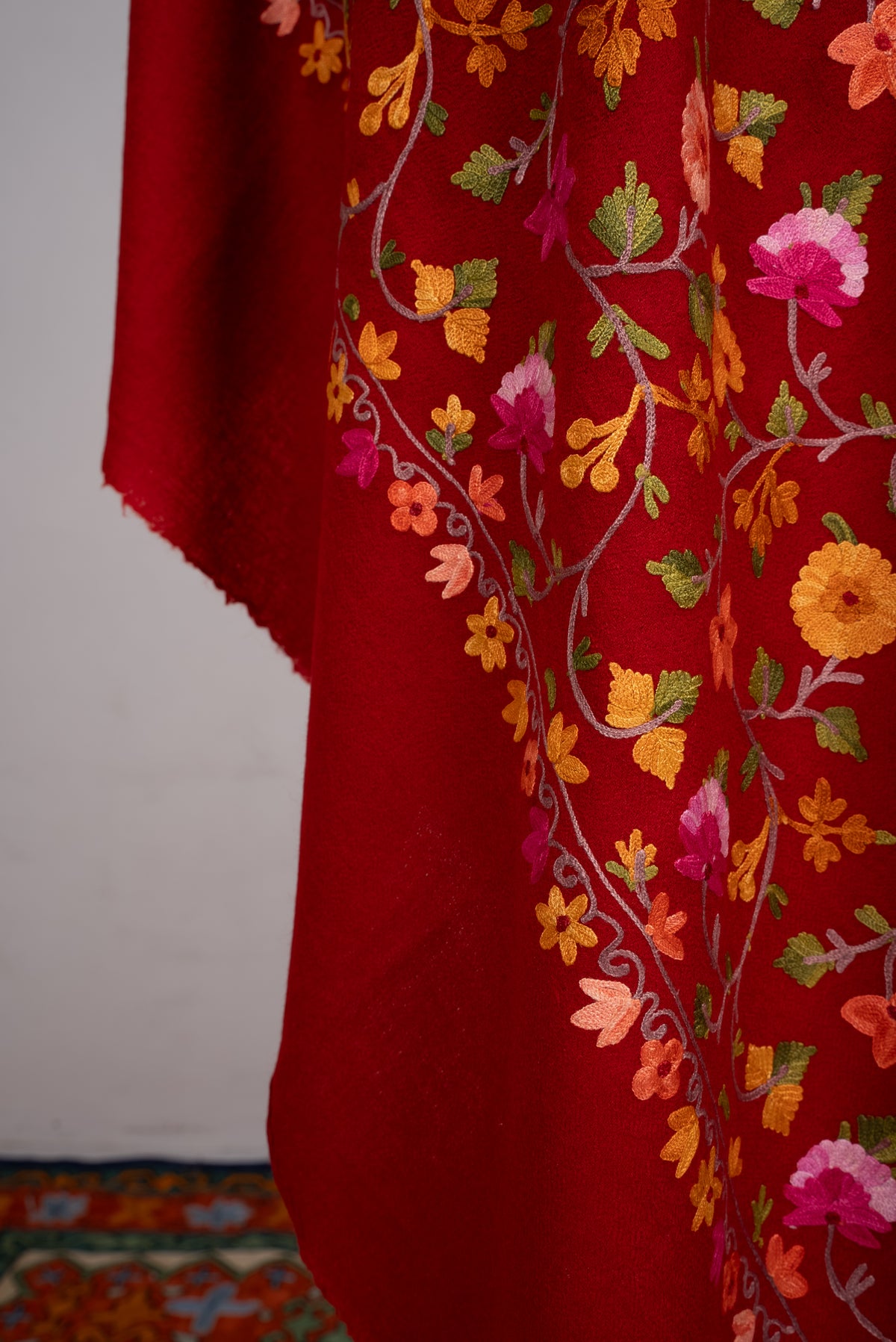 Lower draped detail of the crimson shawl, with colourful Aari floral vines above a narrow border and plain red end
