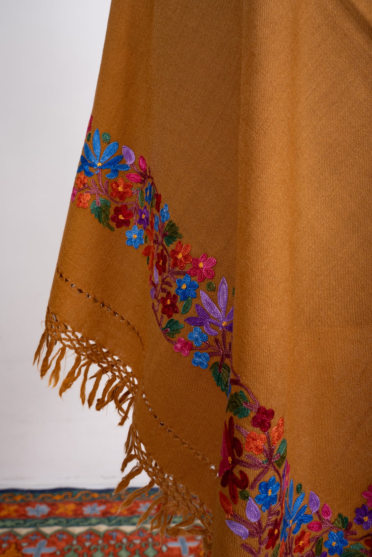 Lower draped corner of the mustard shawl, with bright blue and pink Aari flowers above matching knotted fringe