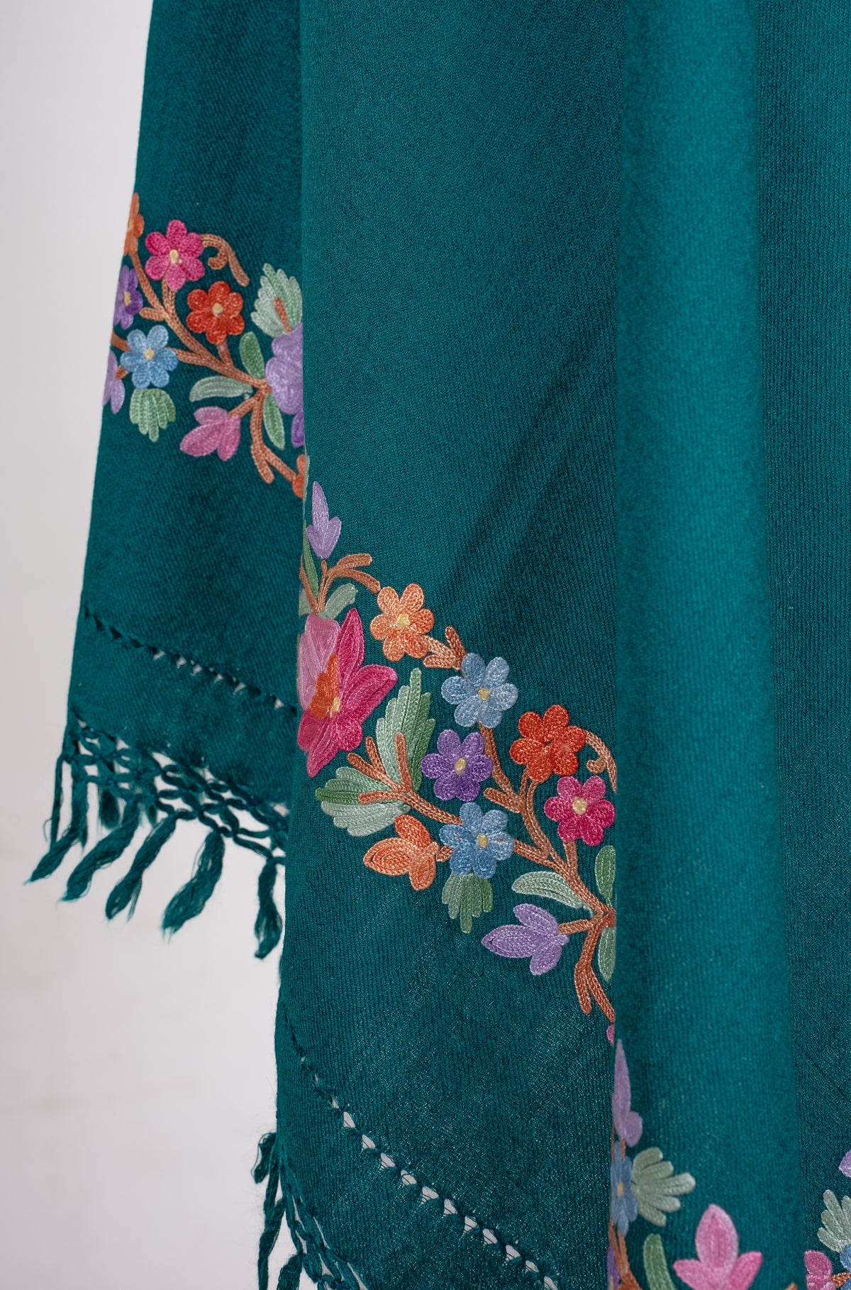 Close-up of a draped teal shawl end, showing pastel Aari flowers along the edge and matching knotted fringe.