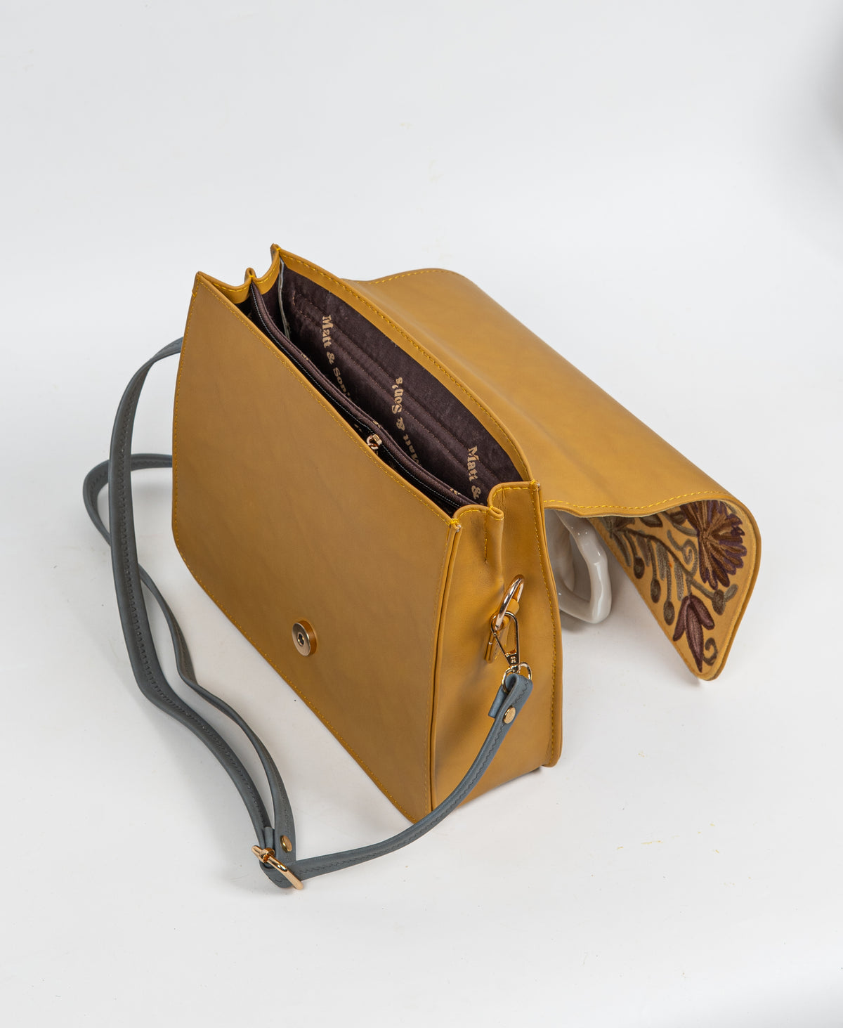 Open handbag with the embroidered flap lifted, revealing dark patterned lining, interior compartments and a round magnetic clasp.