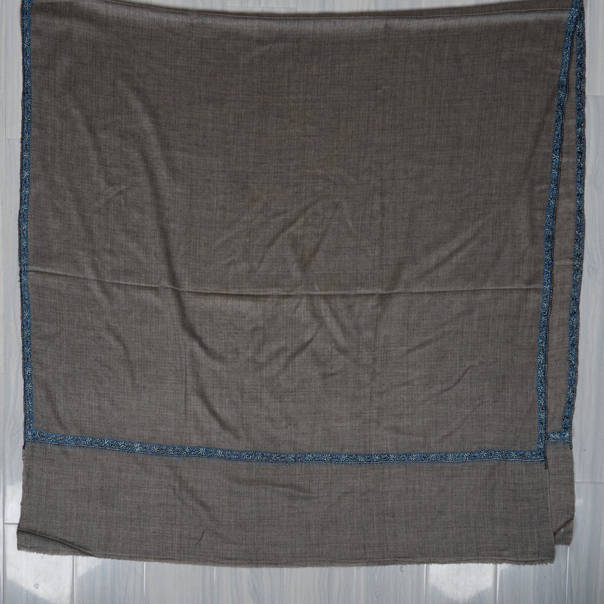 Grey Merino wool shawl laid flat with narrow blue Sozni borders framing a plain centre and end panel.