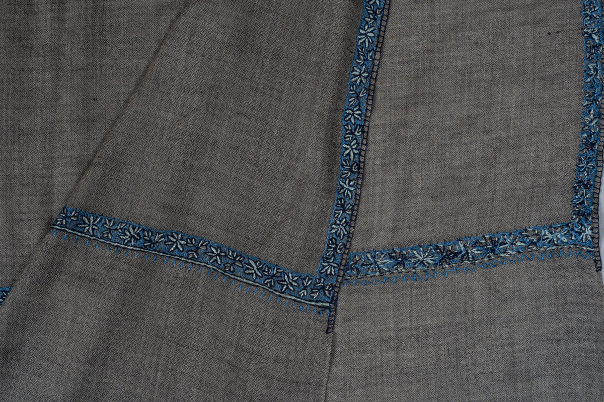 Overlapping corners showing the front and reverse of blue Sozni border stitching on a grey Merino wool shawl.