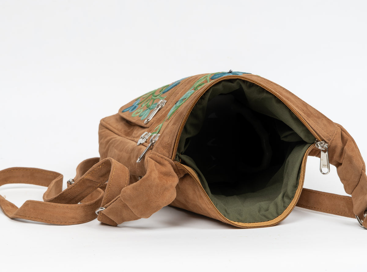 Brown sling bag shown open, revealing a dark olive lining beneath its top zip and floral-embroidered front