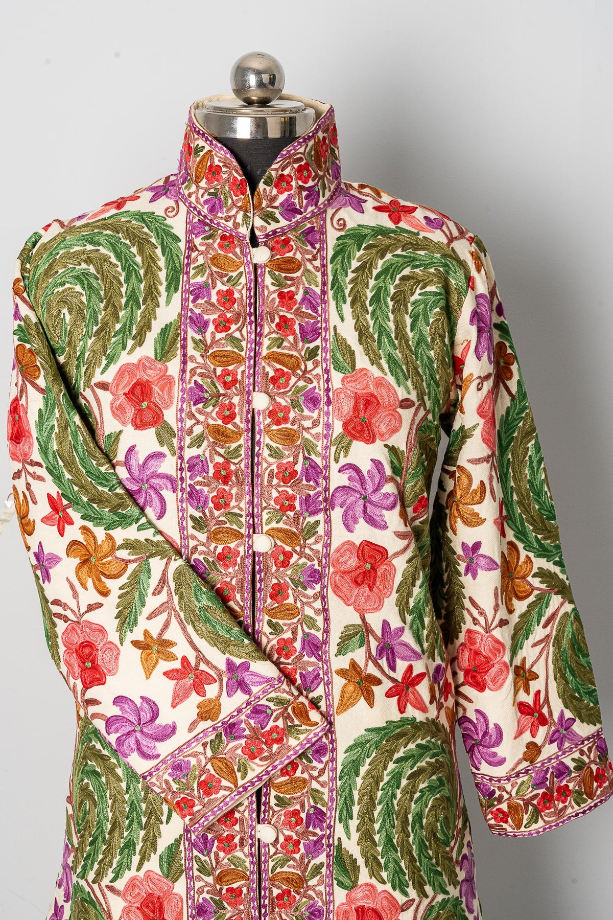 Upper front of the white jacket, showing floral Aari embroidery across the stand collar, chest and folded sleeve